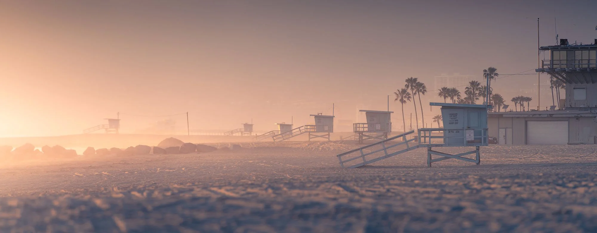 Empty lifeguard towers and palm trees on a sandy beach during sunrise, with haze and a soft glow in the sky.