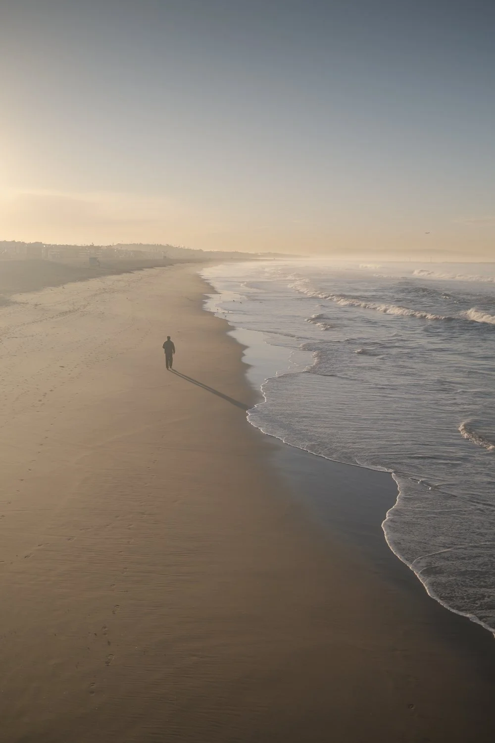 A person walking alone on a sandy beach during sunset or sunrise, with gentle waves crashing onto the shore and a distant city skyline in the background.