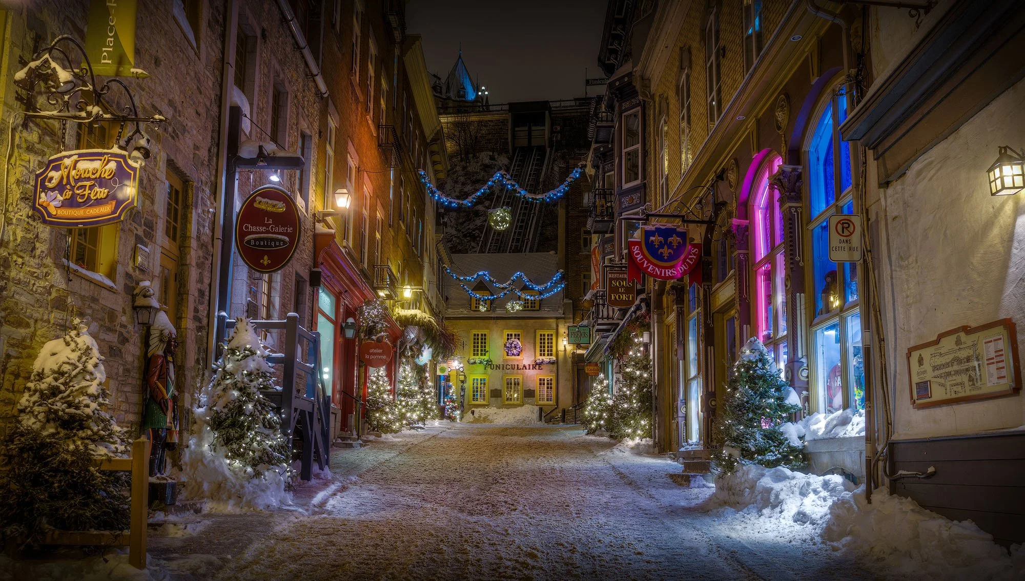 Snow-covered quaint street at night decorated with Christmas lights, small Christmas trees with ornaments, and colorful storefronts illuminated.