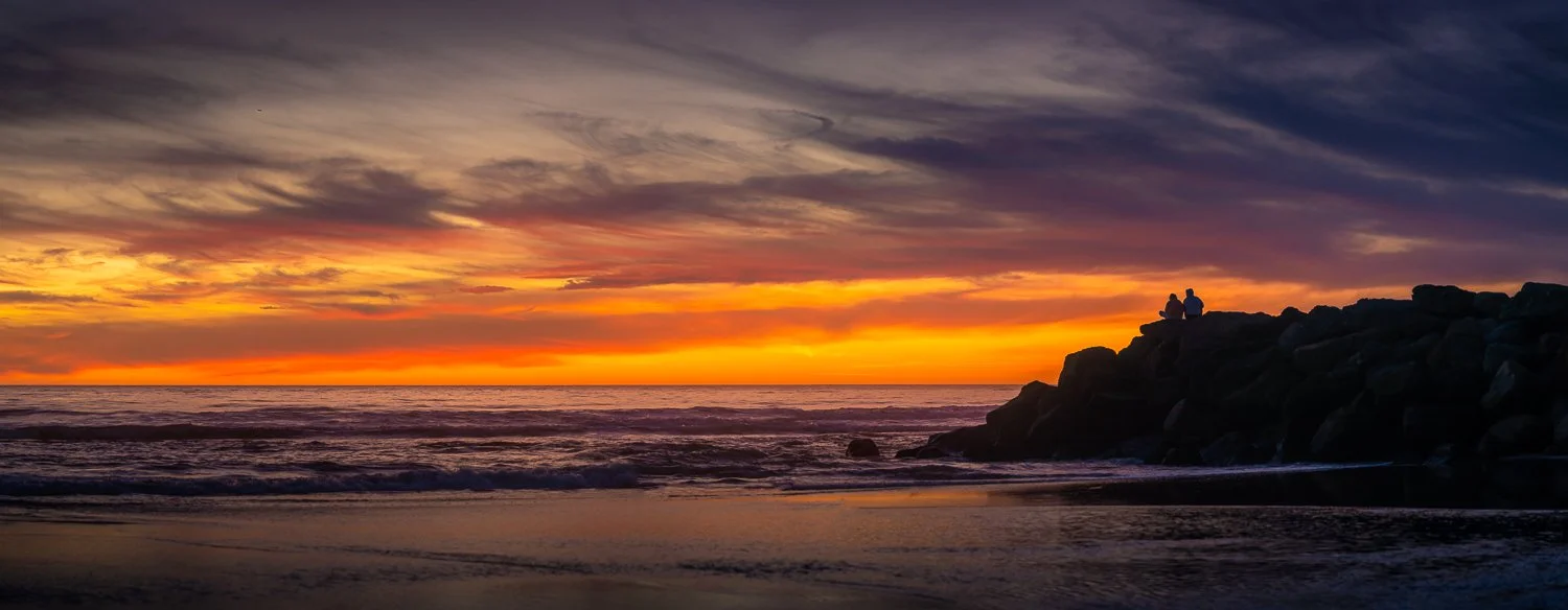 Two people sitting on rocks at sunset on the beach, with colorful sky and ocean waves.