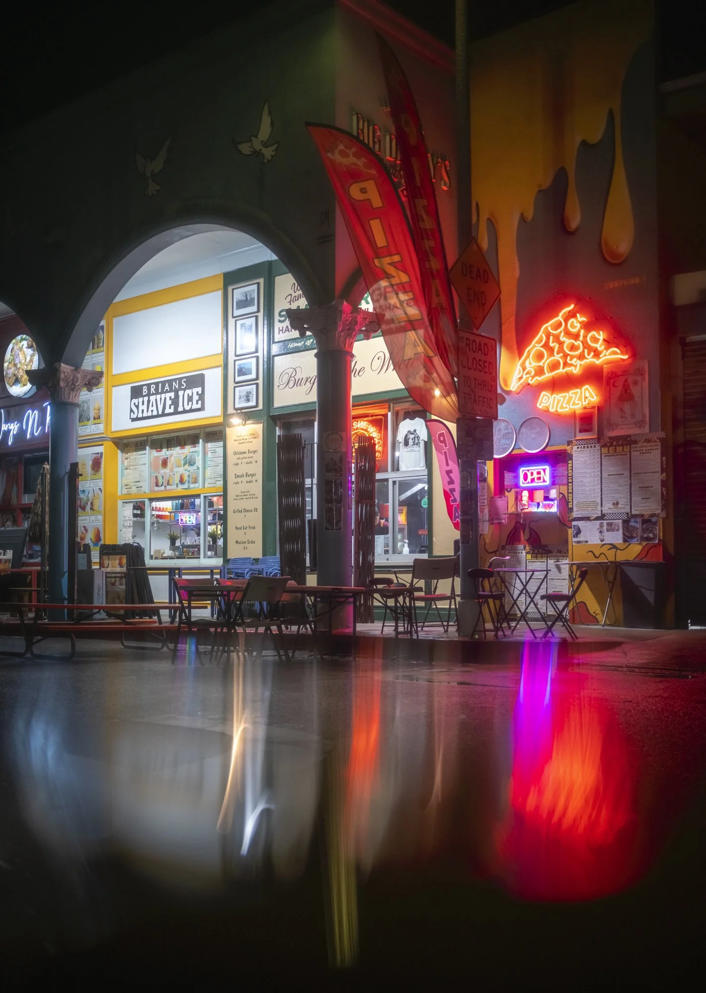Nighttime view of a neon-lit restaurant with neon signs including pizza and open, with empty tables and chairs outside under brightly colored signs and posters.