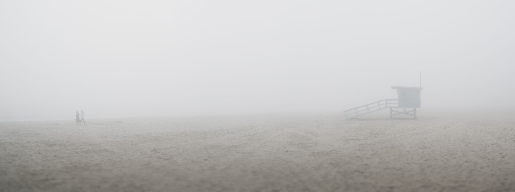 A foggy beach scene with a lifeguard tower on the right and two people walking in the distance on the left.