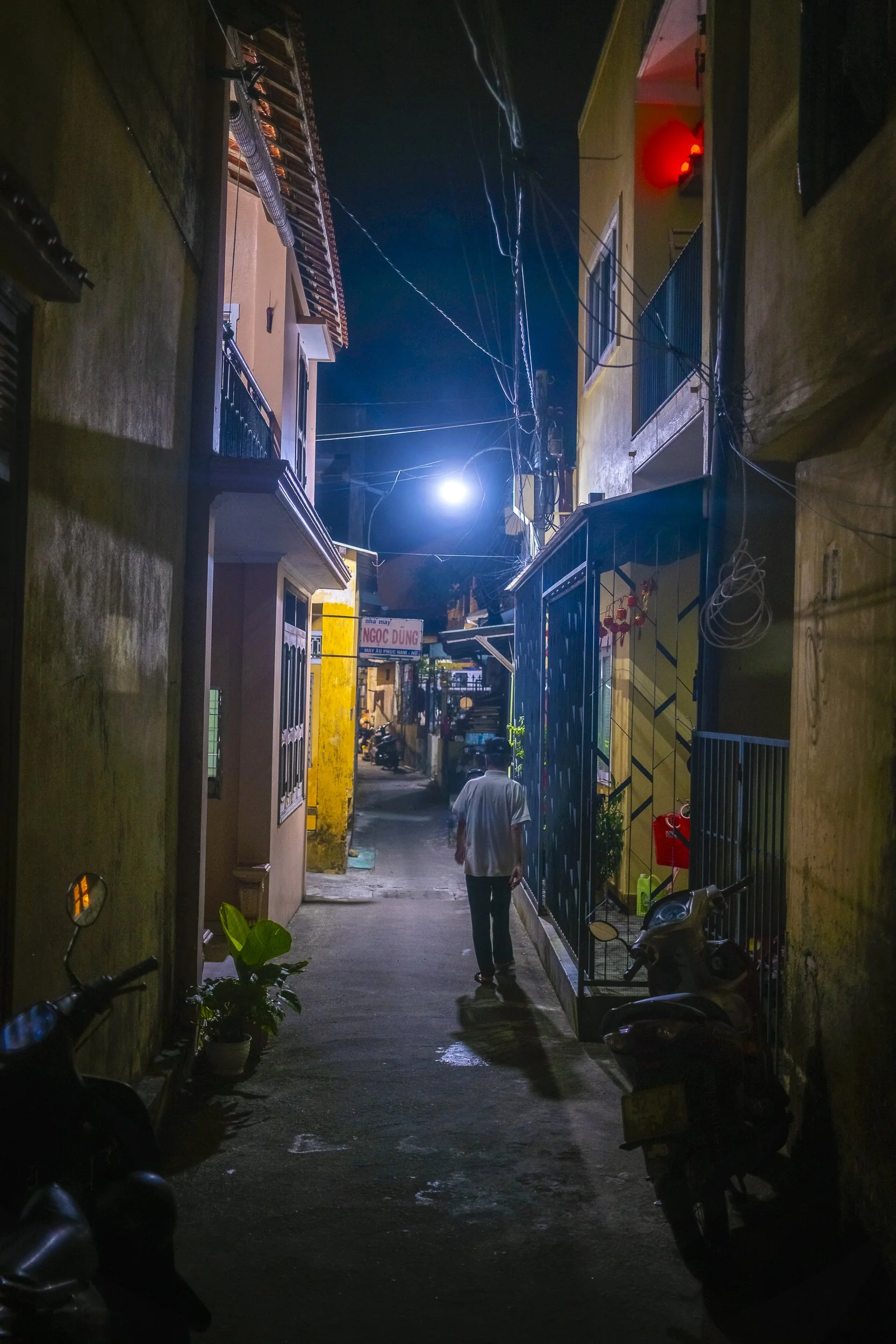 A narrow alley at night illuminated by a bright streetlight, with a man walking away from the camera, parked scooters on either side, colorful houses, and electrical wires overhead.