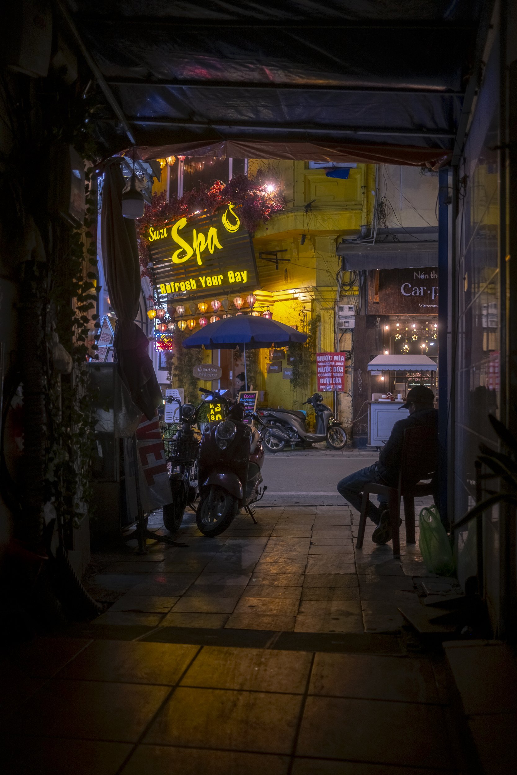Night scene of a narrow street view through an open doorway, with a person sitting under an umbrella, motorcycles parked on the sidewalk, illuminated signs, and colorful hanging lanterns.