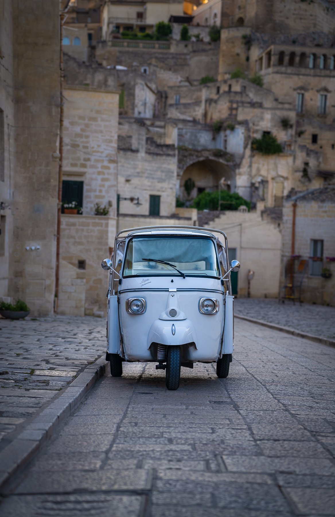 A small, vintage white three-wheeled vehicle parked on a cobblestone street in front of stone buildings with a hillside in the background.