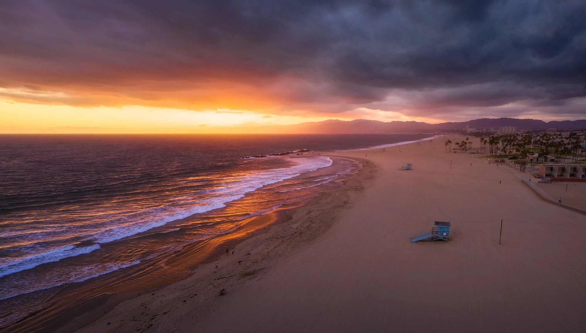 Empty beach at sunset with dark clouds overhead and distant mountains in the background.