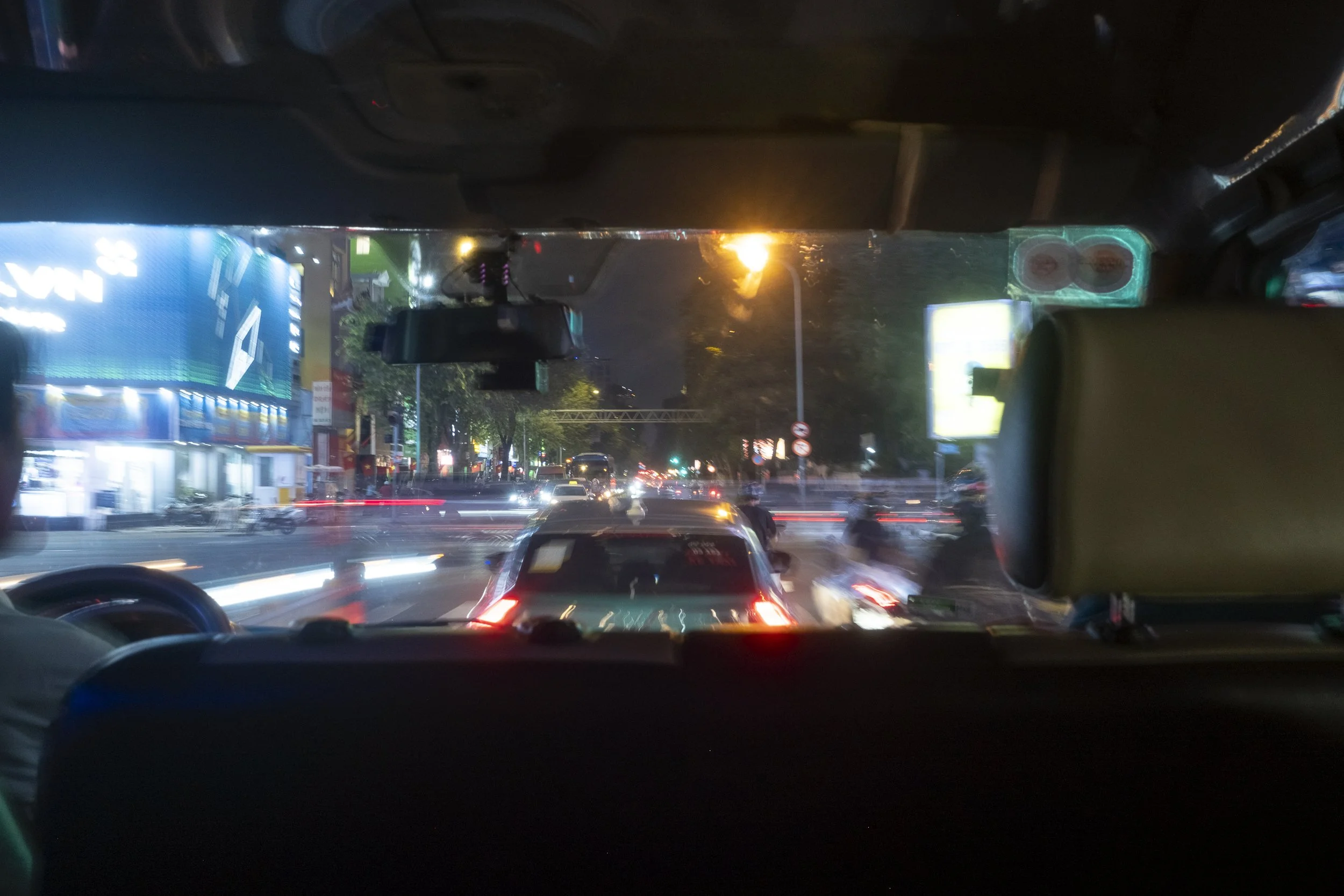 Nighttime view from inside a vehicle showing a busy city street with cars, blurred lights, and buildings with illuminated signs.