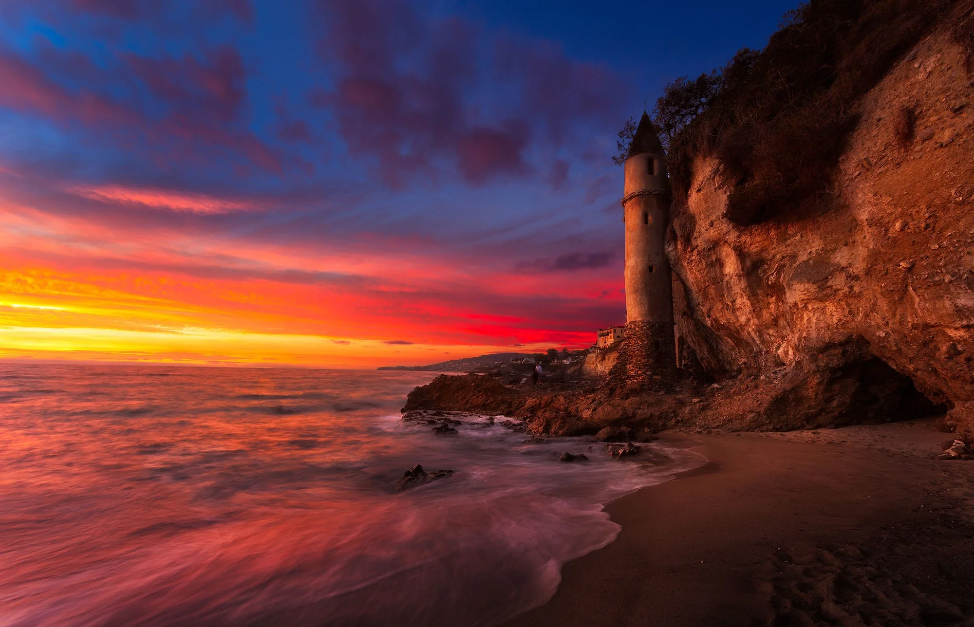 A sunset over the ocean with a tower built into a rocky cliff, colorful clouds, and waves crashing on the shore.