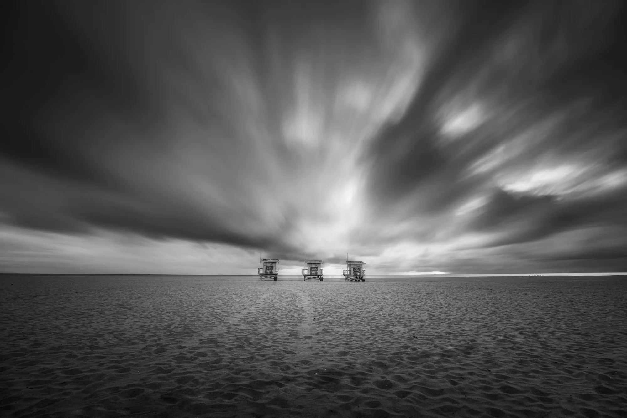 Black and white photo of a beach with three lifeguard towers in the distance and dramatic clouds in the sky.