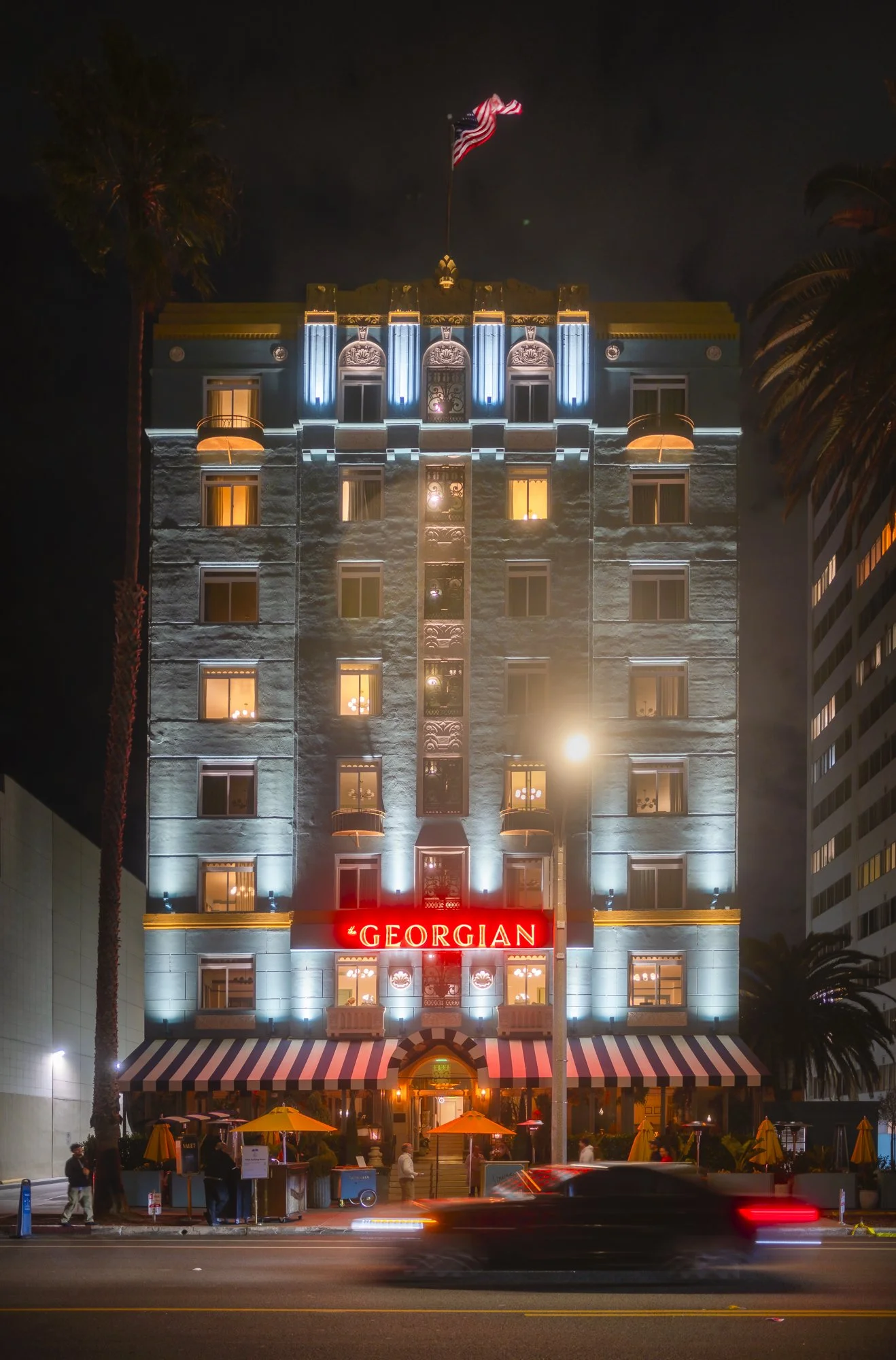 Night view of the Georgian hotel building illuminated with blue and white lights, with a red neon sign reading 'Georgian' at the entrance, and people walking outside under yellow umbrellas.