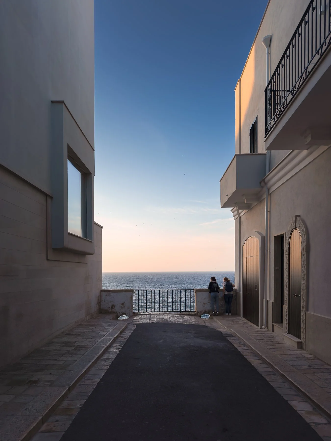 Two people stand on a balcony overlooking the ocean at sunset, framed by white buildings on either side.