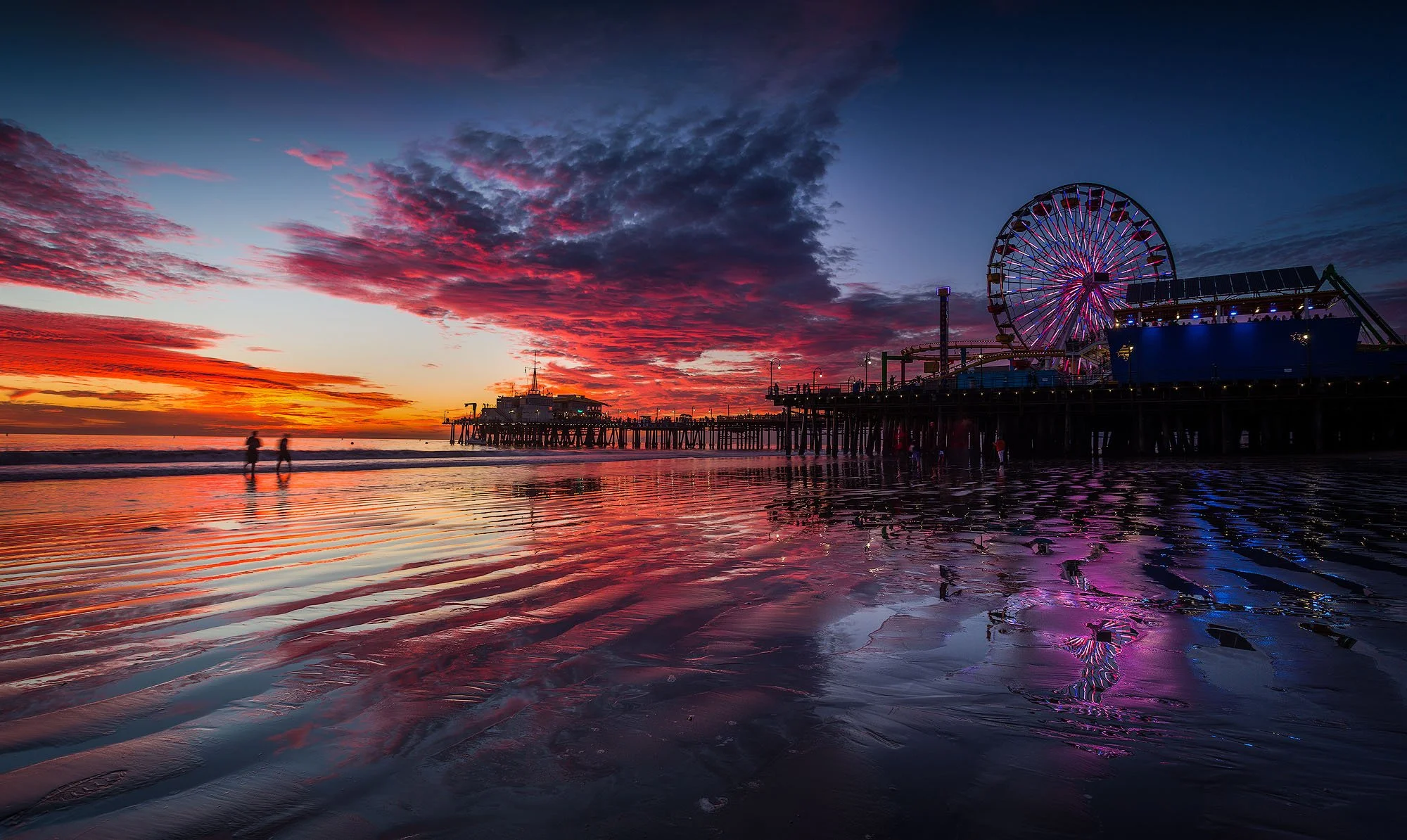Sunset over a pier with a ferris wheel and amusement park rides, reflected in the wet sand with a couple walking along the beach.