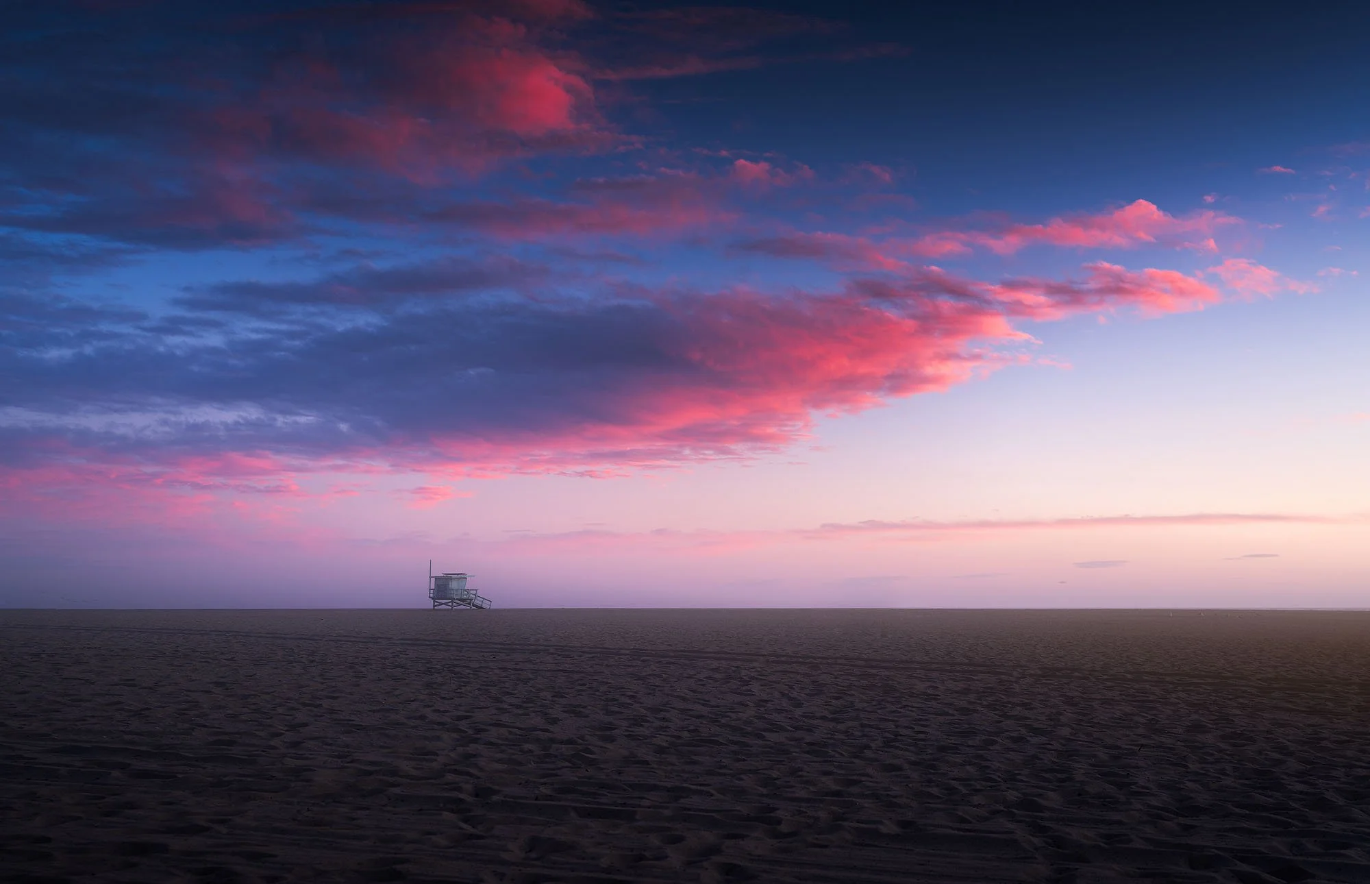 A beach scene at sunset with pink and purple clouds and an empty lifeguard tower in the distance.