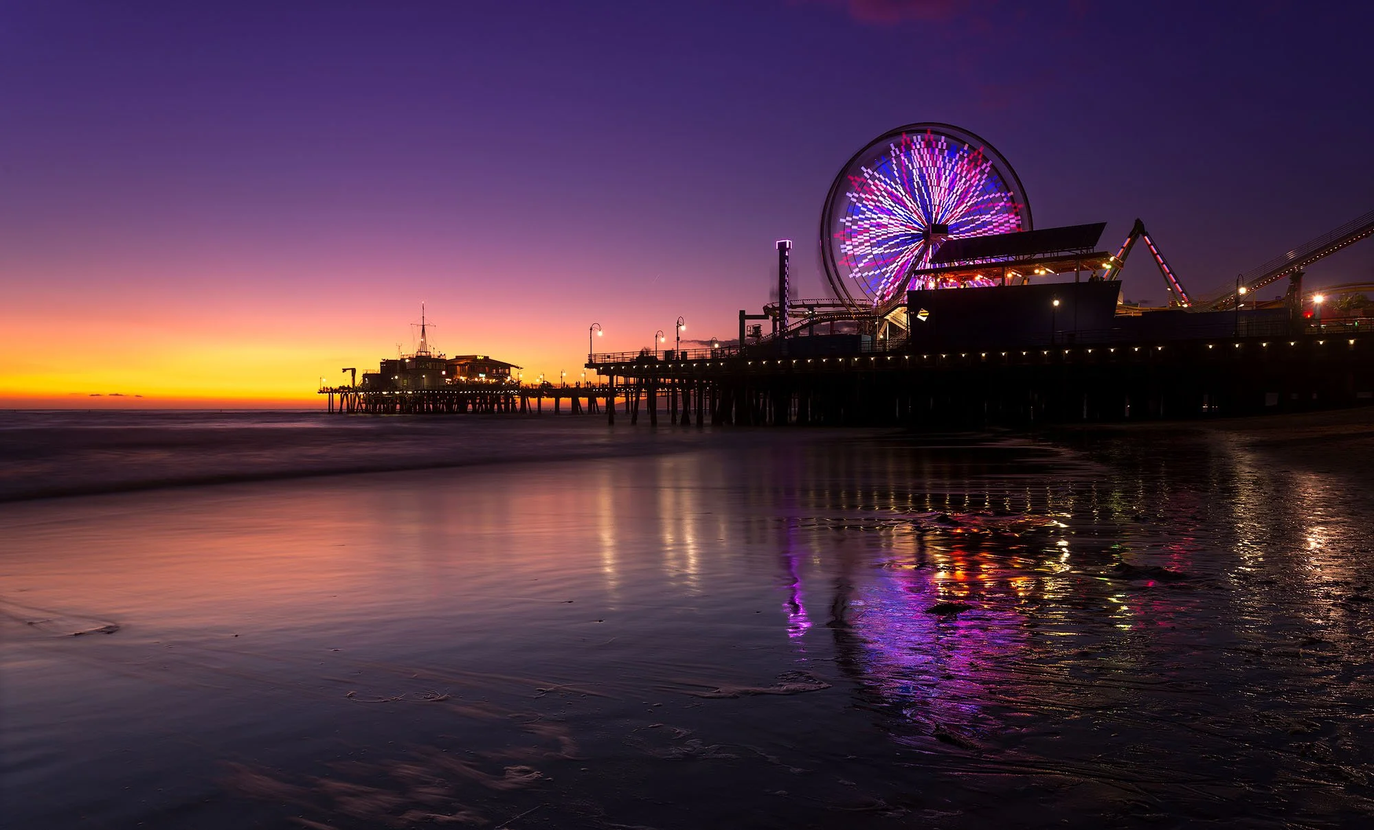 Sunset over a pier with a large illuminated Ferris wheel and reflection in the water nearby in Santa Monica, California.