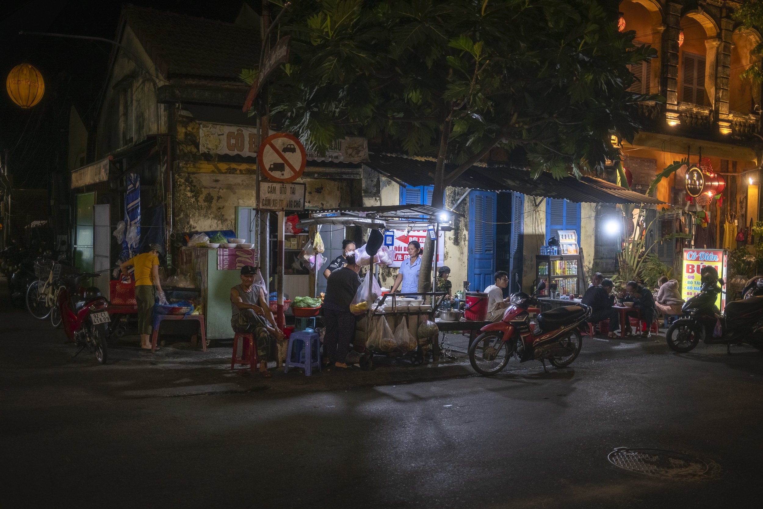 Night street scene with street vendors, small eatery, and people sitting at tables, illuminated by streetlights and shop lights, with motorcycles parked on the side.