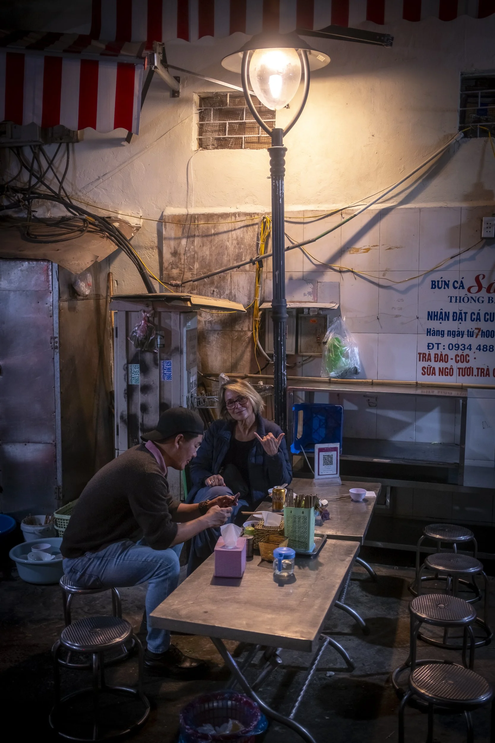 Two people sitting at a small table outdoors next to a street lamp at night, with one person gesturing and the other looking at their phone. The scene includes stools, a tissue box, and various items on the table against a background of a wall with w