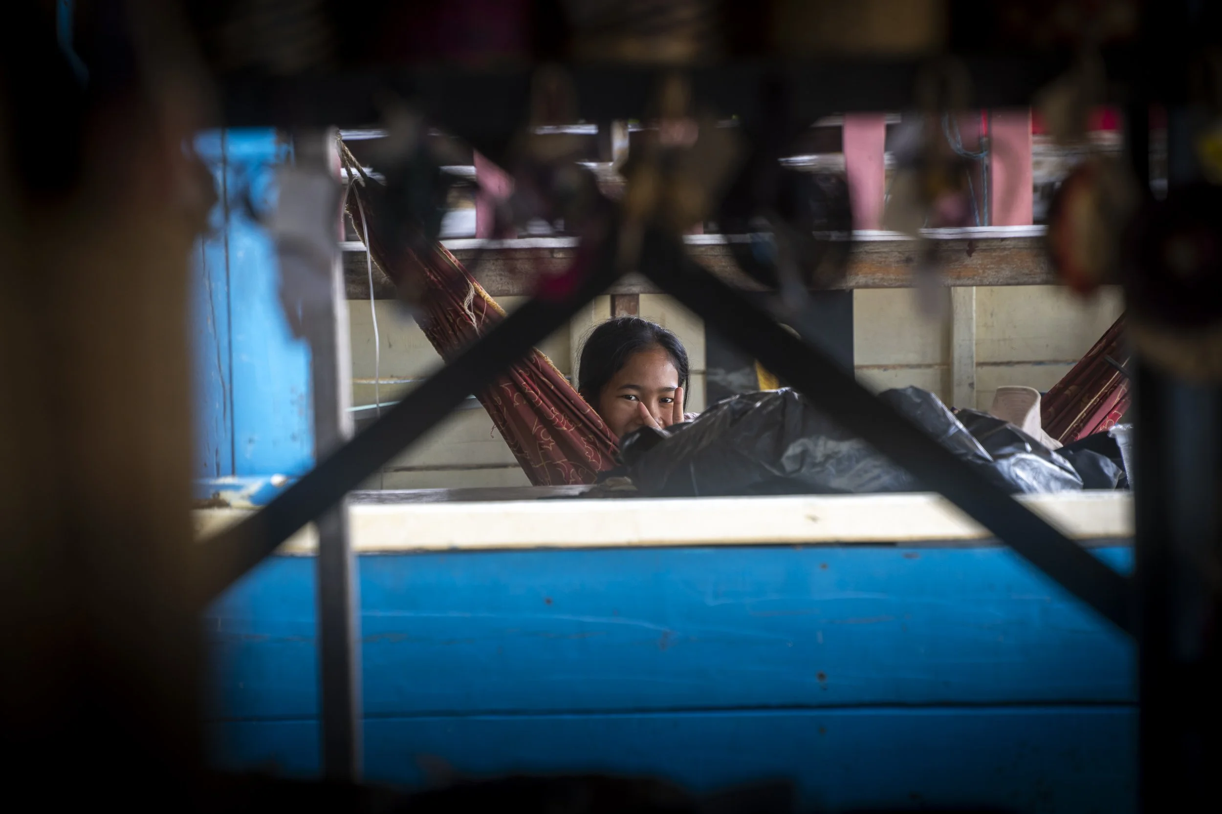 A young woman lying in a hammock, smiling and peeking over a black plastic bag, seen through a metal grate.