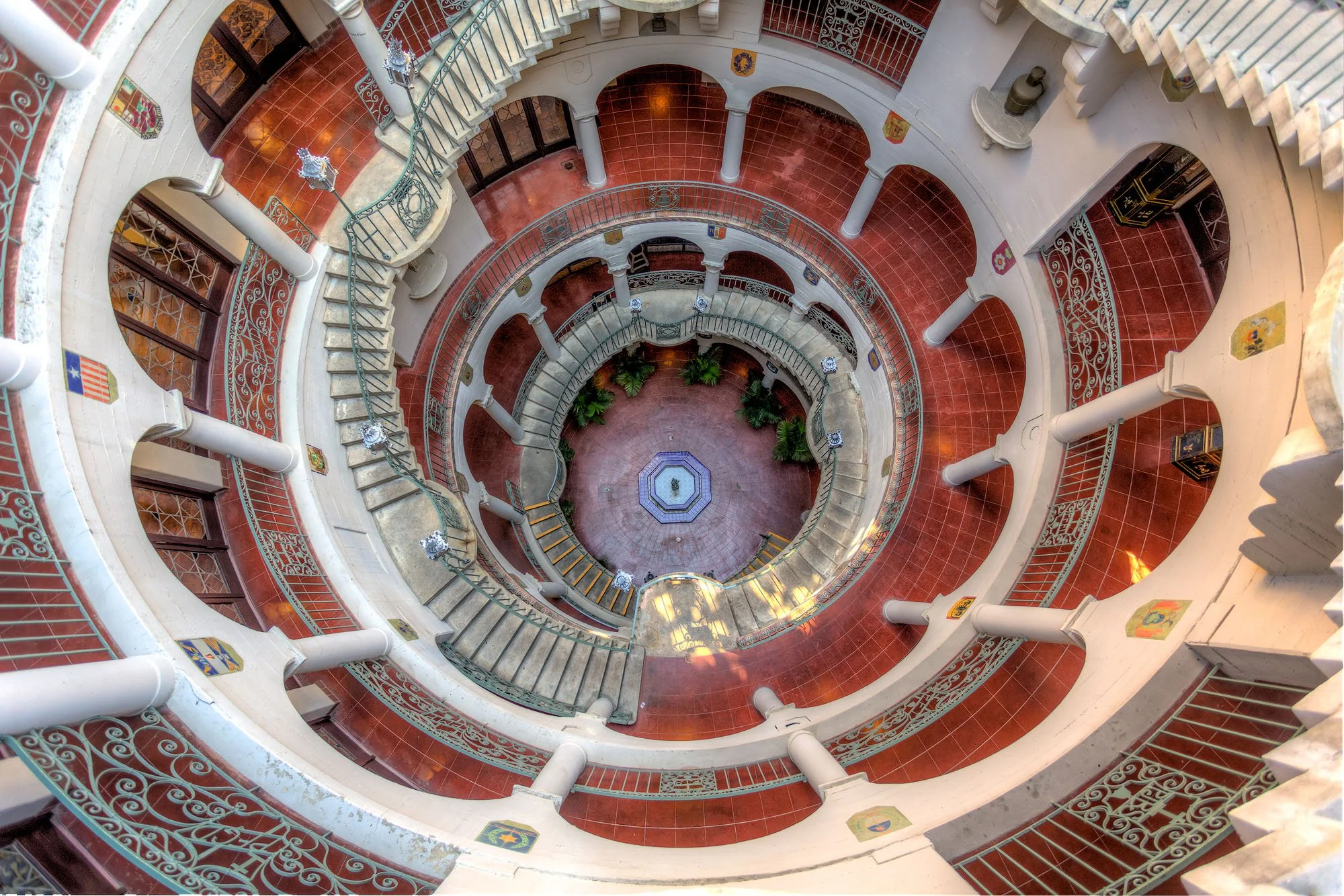 A multi-story atrium with a central fountain, surrounded by white columns, ornate iron railings, and red-tiled floors, viewed from the top.