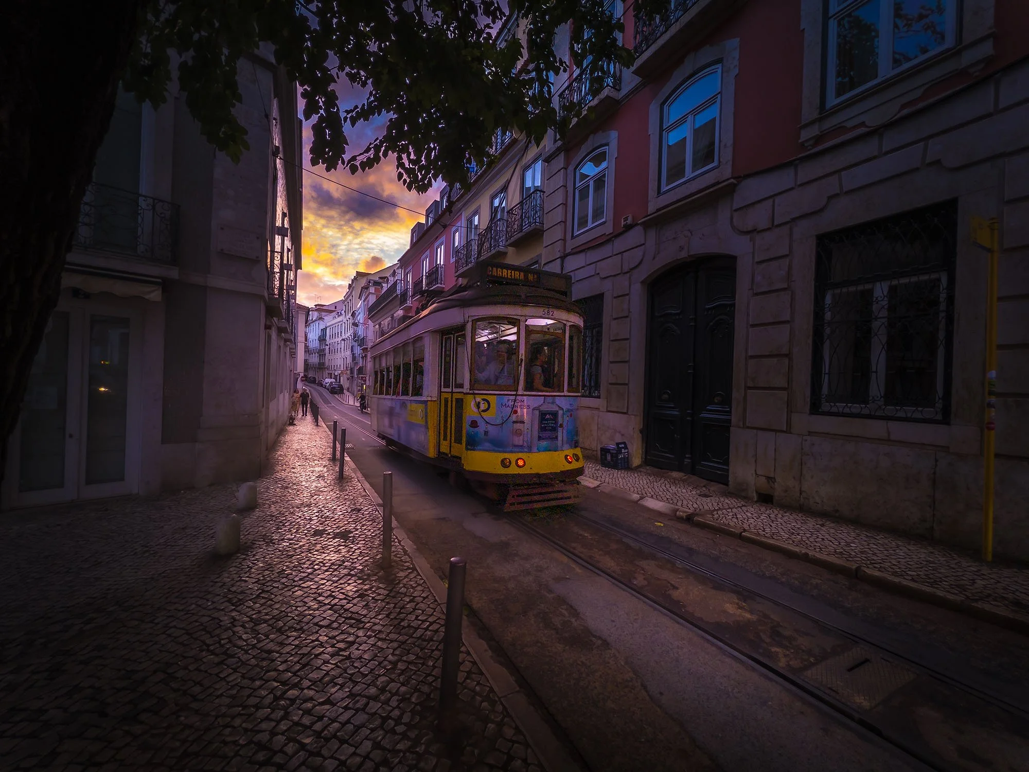 A colorful tram on a narrow street during sunset with buildings on both sides and a tree at the top left.