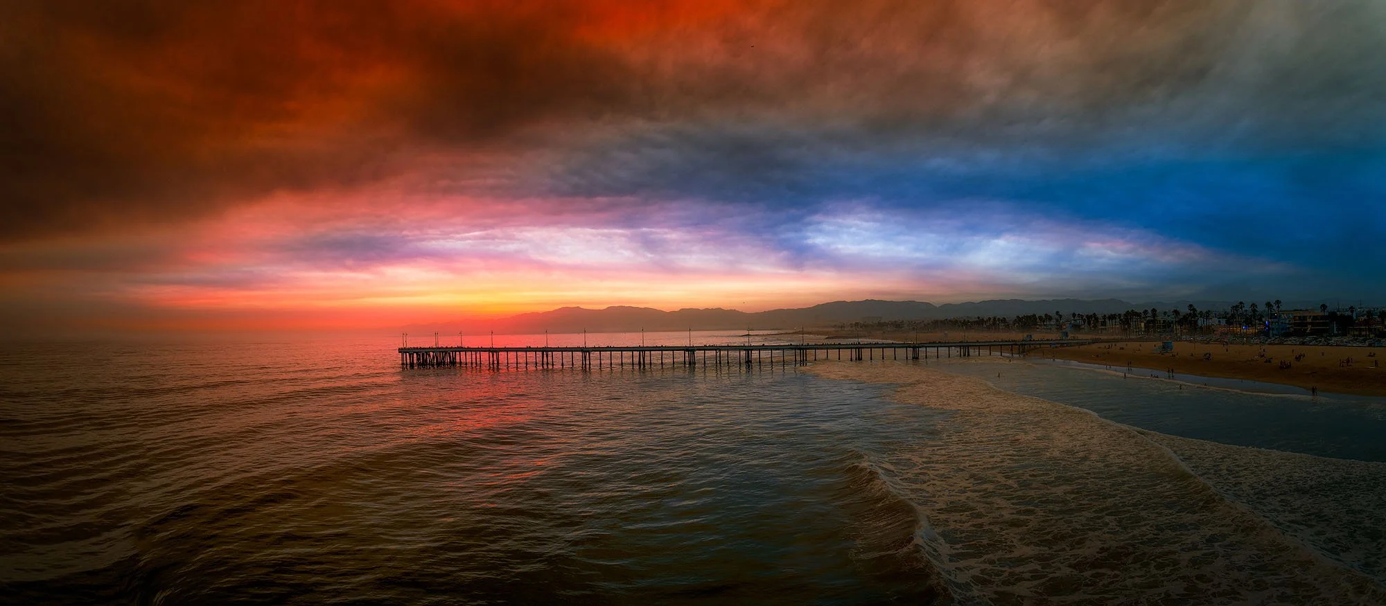 Sunset over the ocean with a pier extending into the water and a beach with people and palm trees in the background, sky filled with colorful clouds.