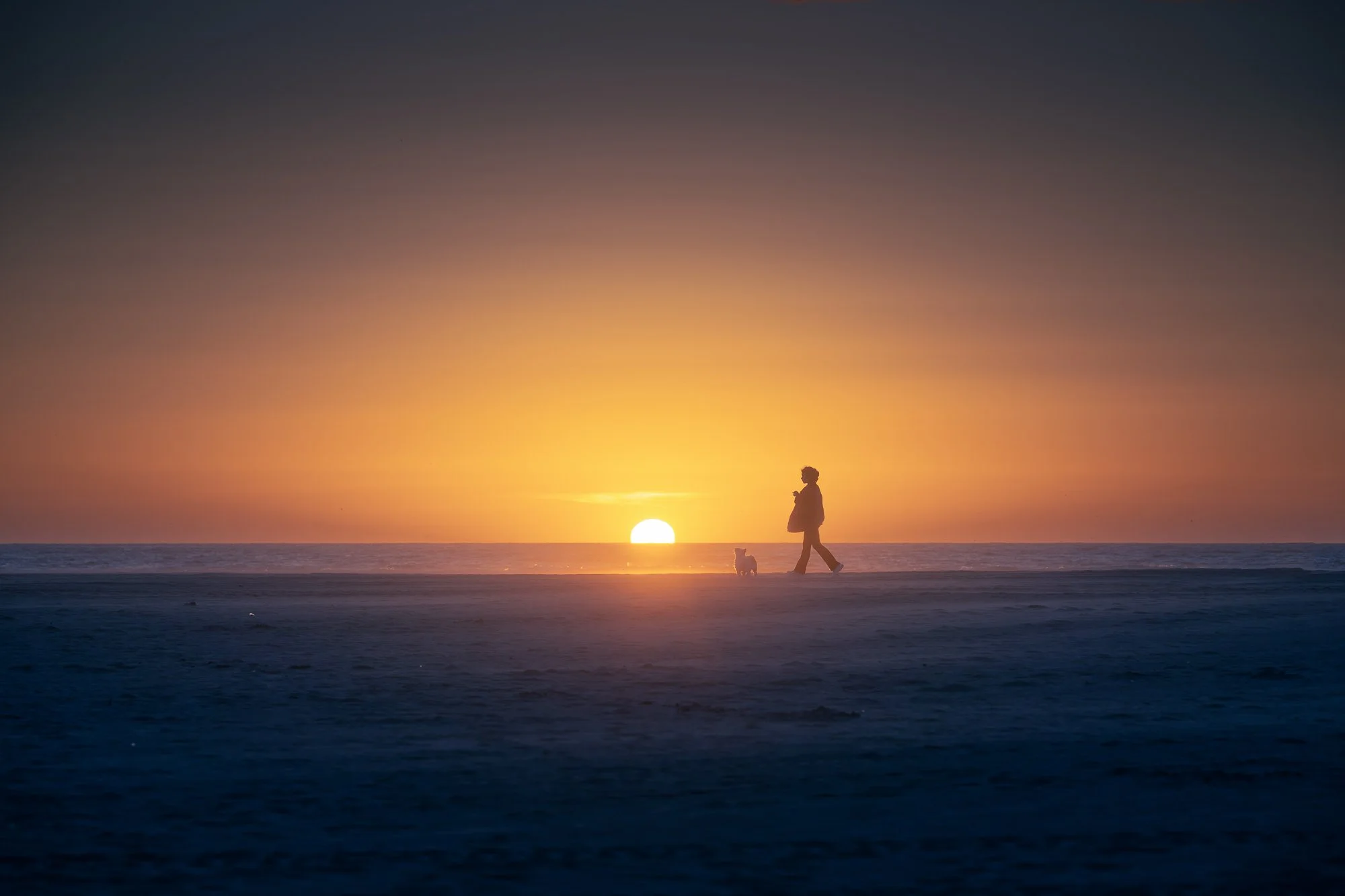 A person walking a dog on a beach during sunset, with the sun near the horizon and the sky displaying warm orange and yellow tones.