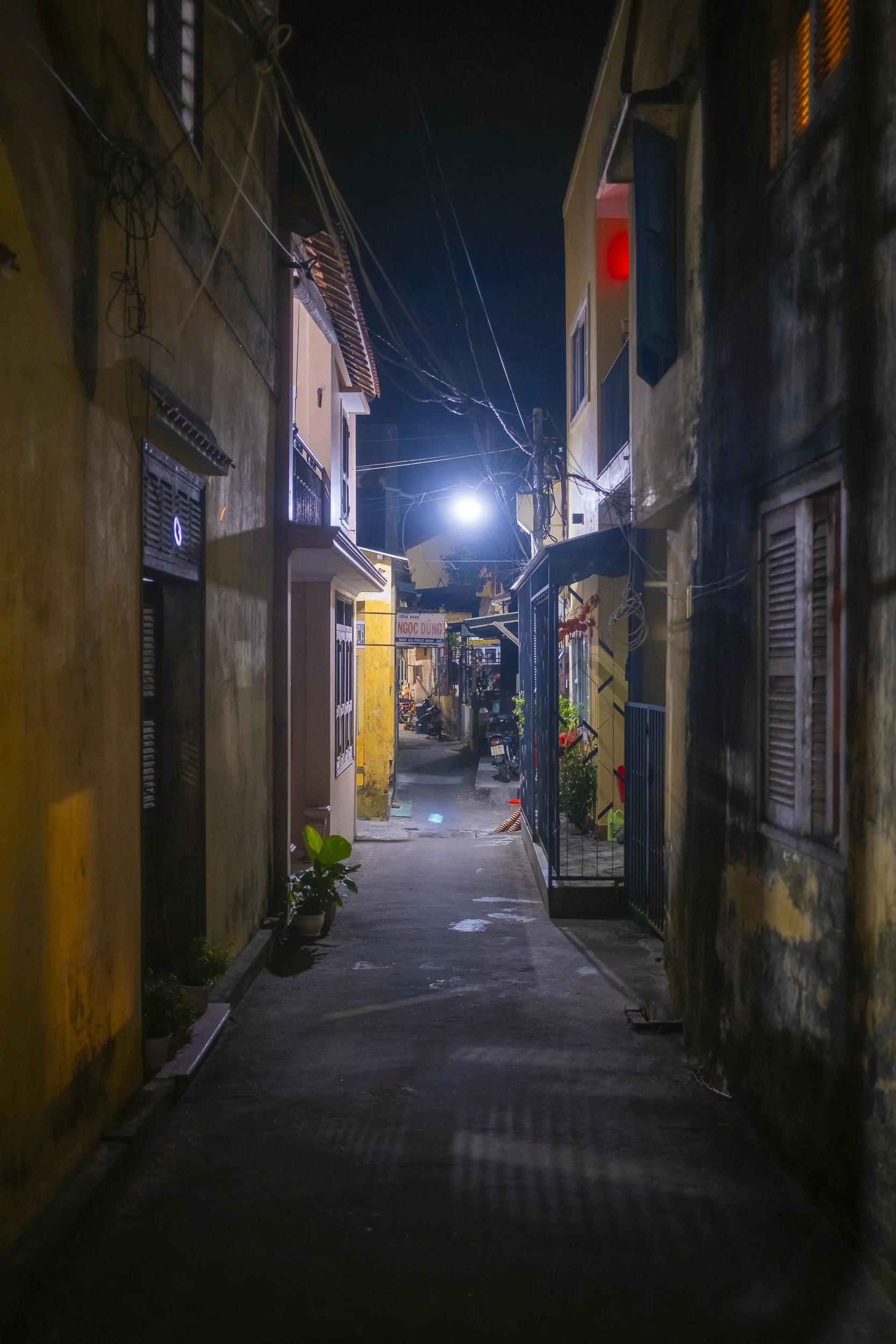Night view of a narrow alleyway between old buildings with potted plants, illuminated by a streetlight, with motorcycles and signs in the background.