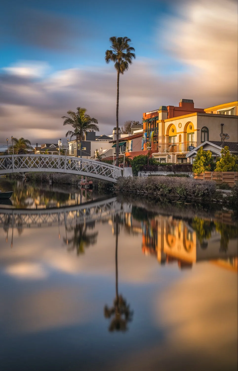 Colorful houses with palm trees along a waterway, with a white bridge and reflections in the water at sunset.