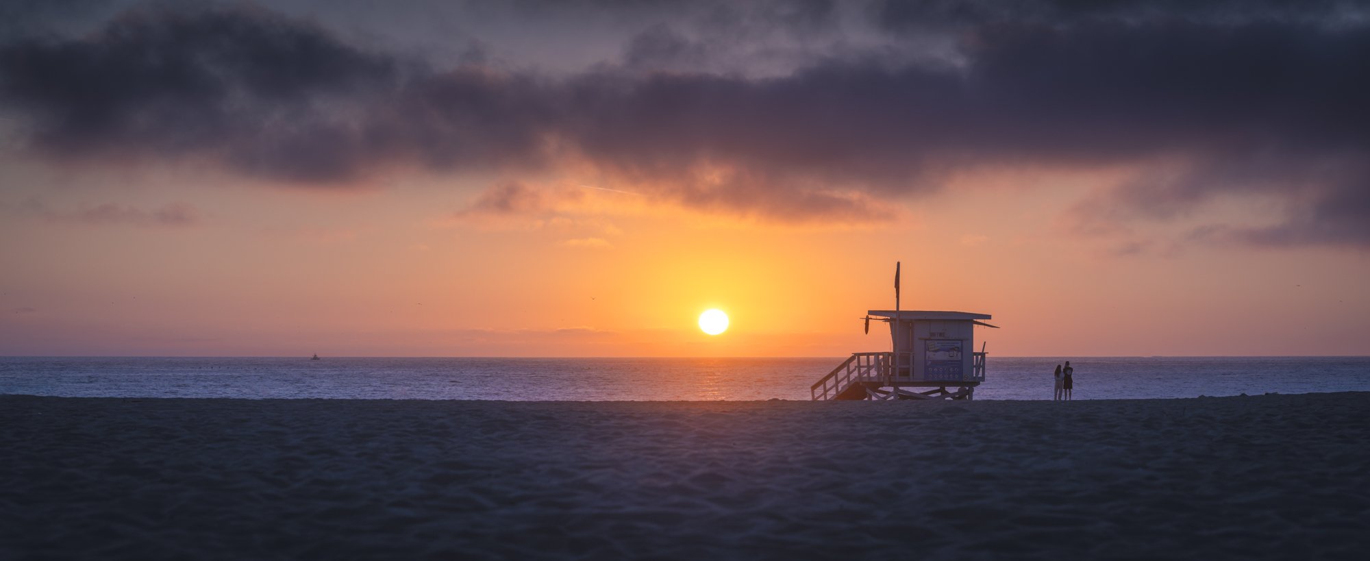Sunset over the ocean with silhouetted lifeguard tower and two people standing on the beach.