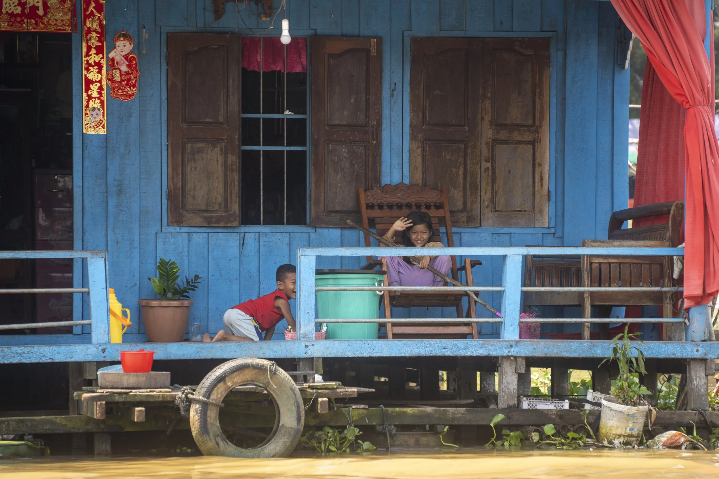 Two children playing on a blue wooden house with a porch near water. The girl is sitting on a wooden chair, waving, while the boy is crouching nearby with a big smile.
