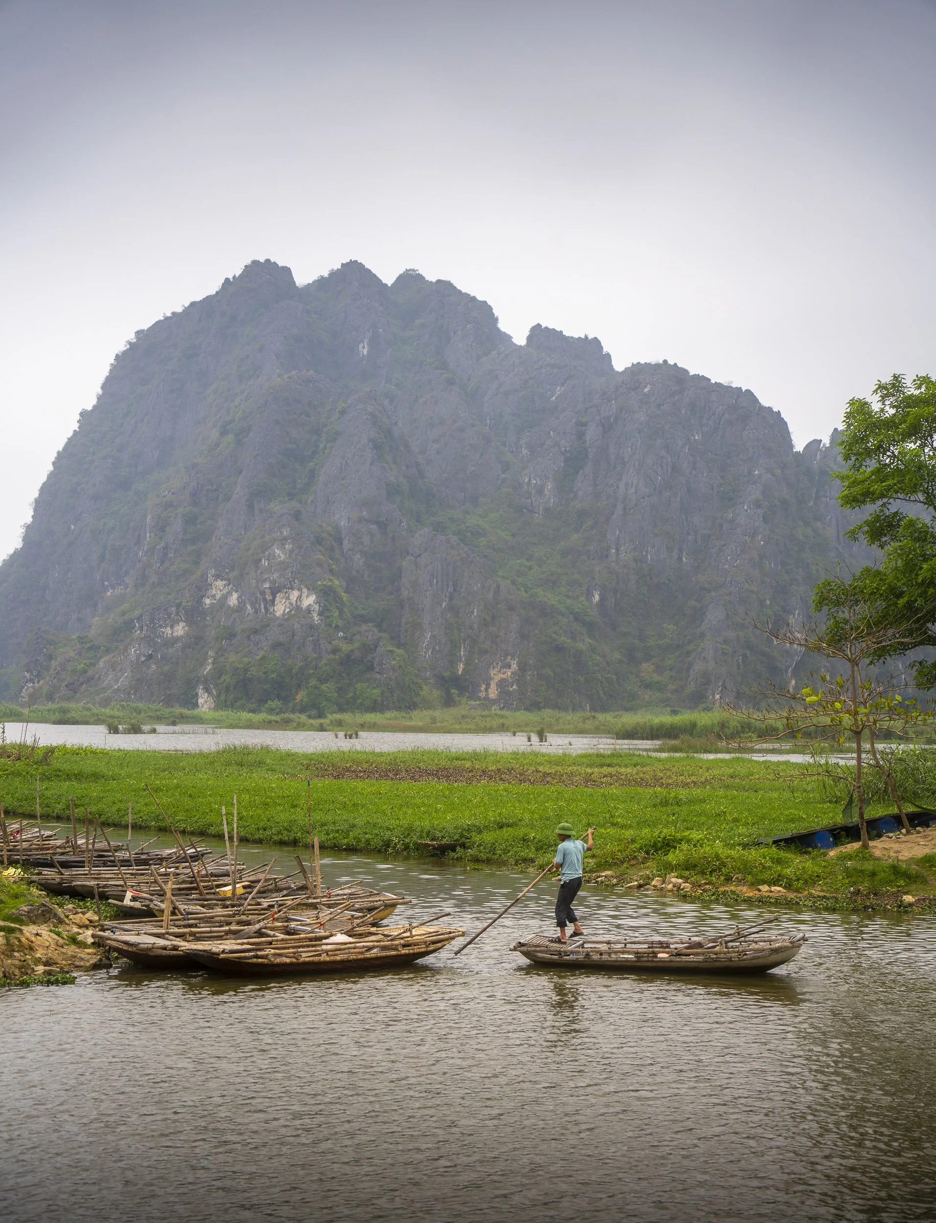A person standing on a bamboo raft on a river, surrounded by lush green landscape with a mountain in the background.