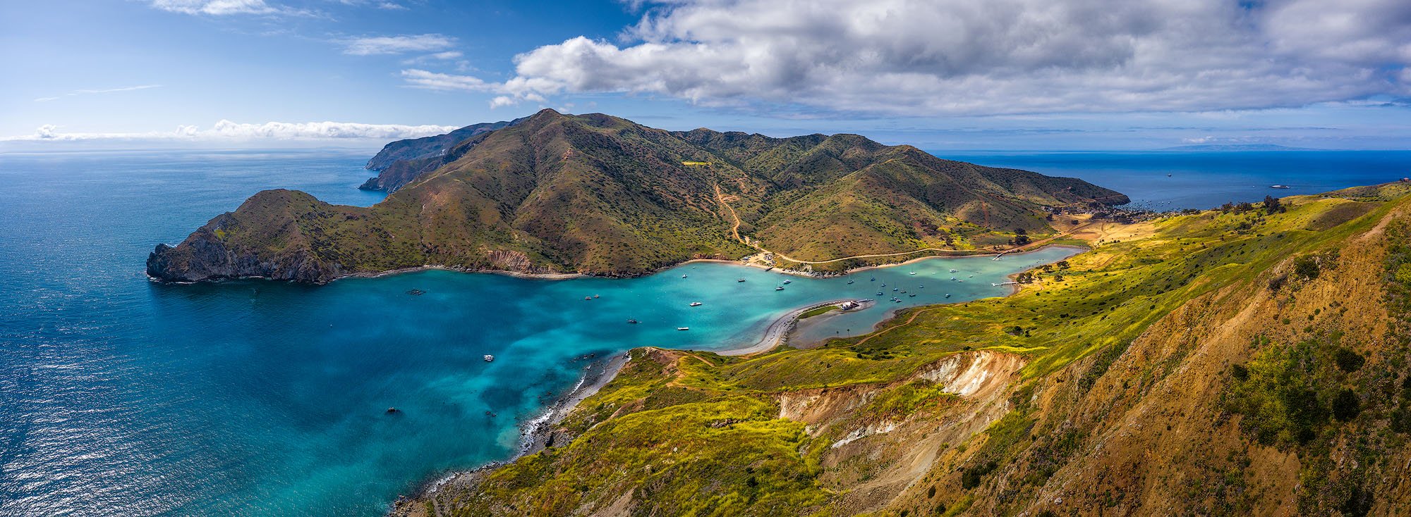 A panoramic view of a coastal bay surrounded by green hills and mountains with boats anchored in the water and a cloudy sky overhead.