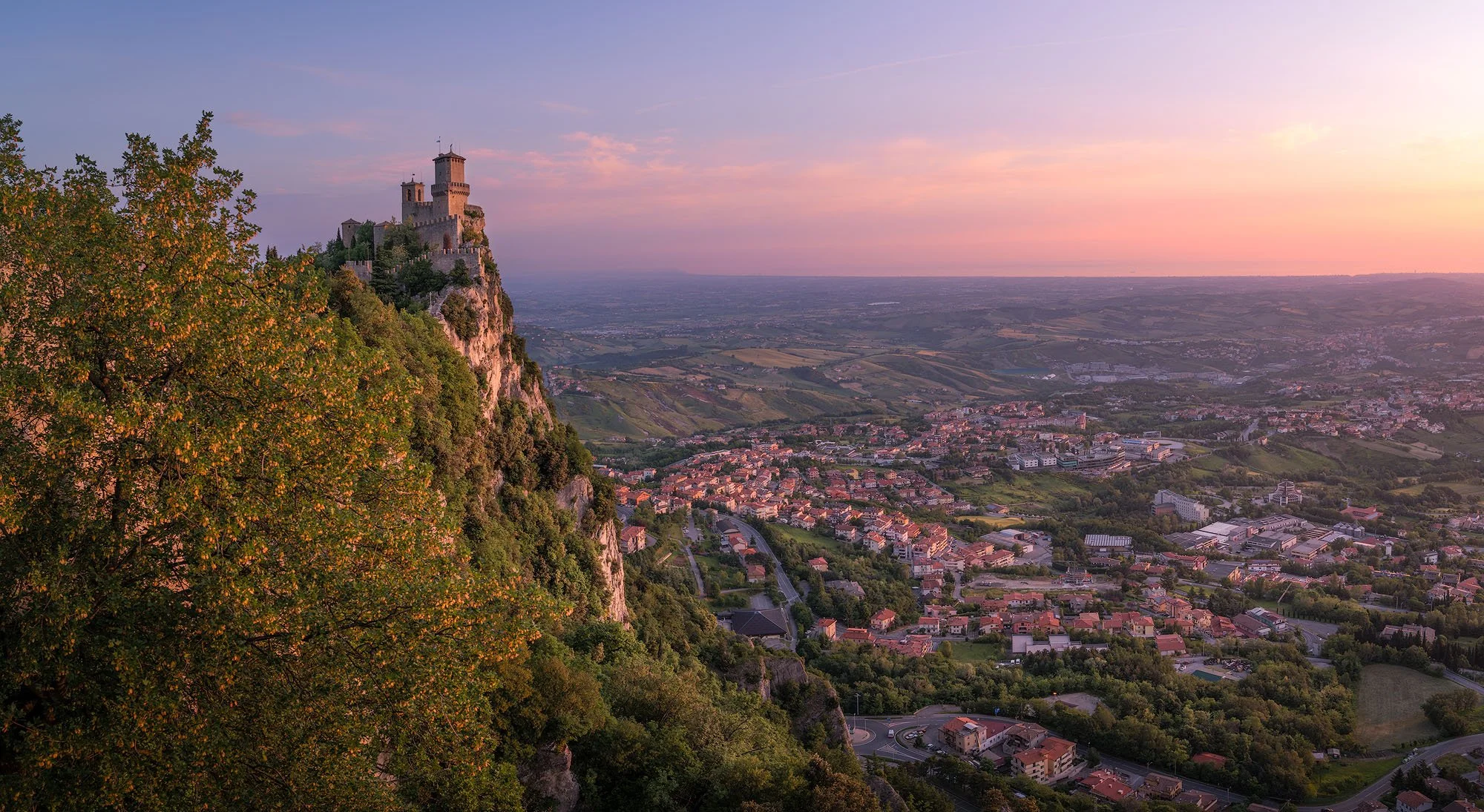 A castle on a rocky hill overlooking a city at sunset, with rolling fields and a distant horizon.