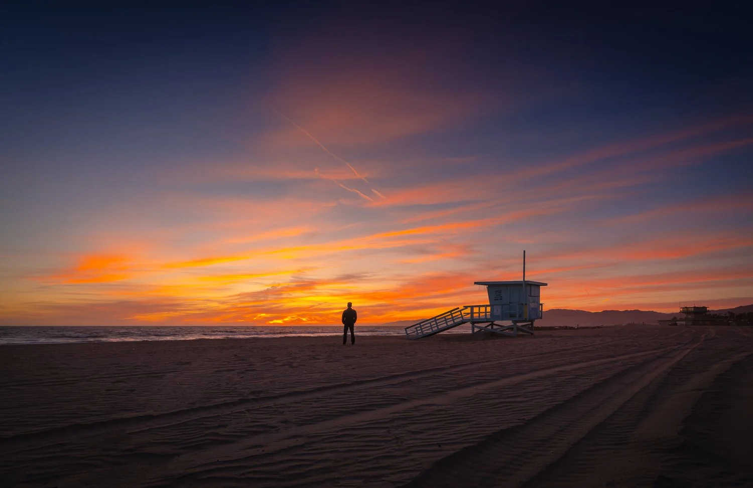 A person standing on a beach at sunset, near a lifeguard tower, with colorful sky and clouds.