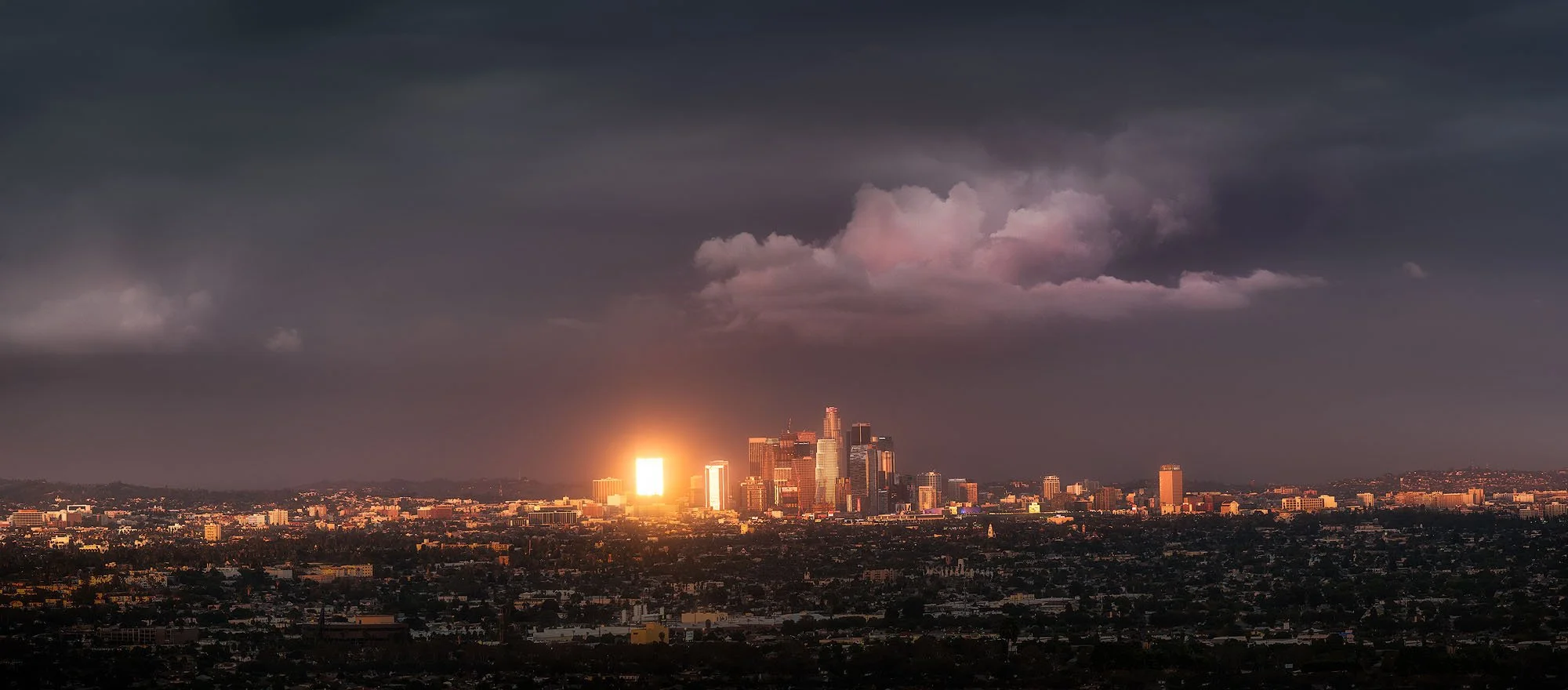 Dark storm clouds over the Los Angeles skyline at sunset, with sunlight reflecting off buildings.
