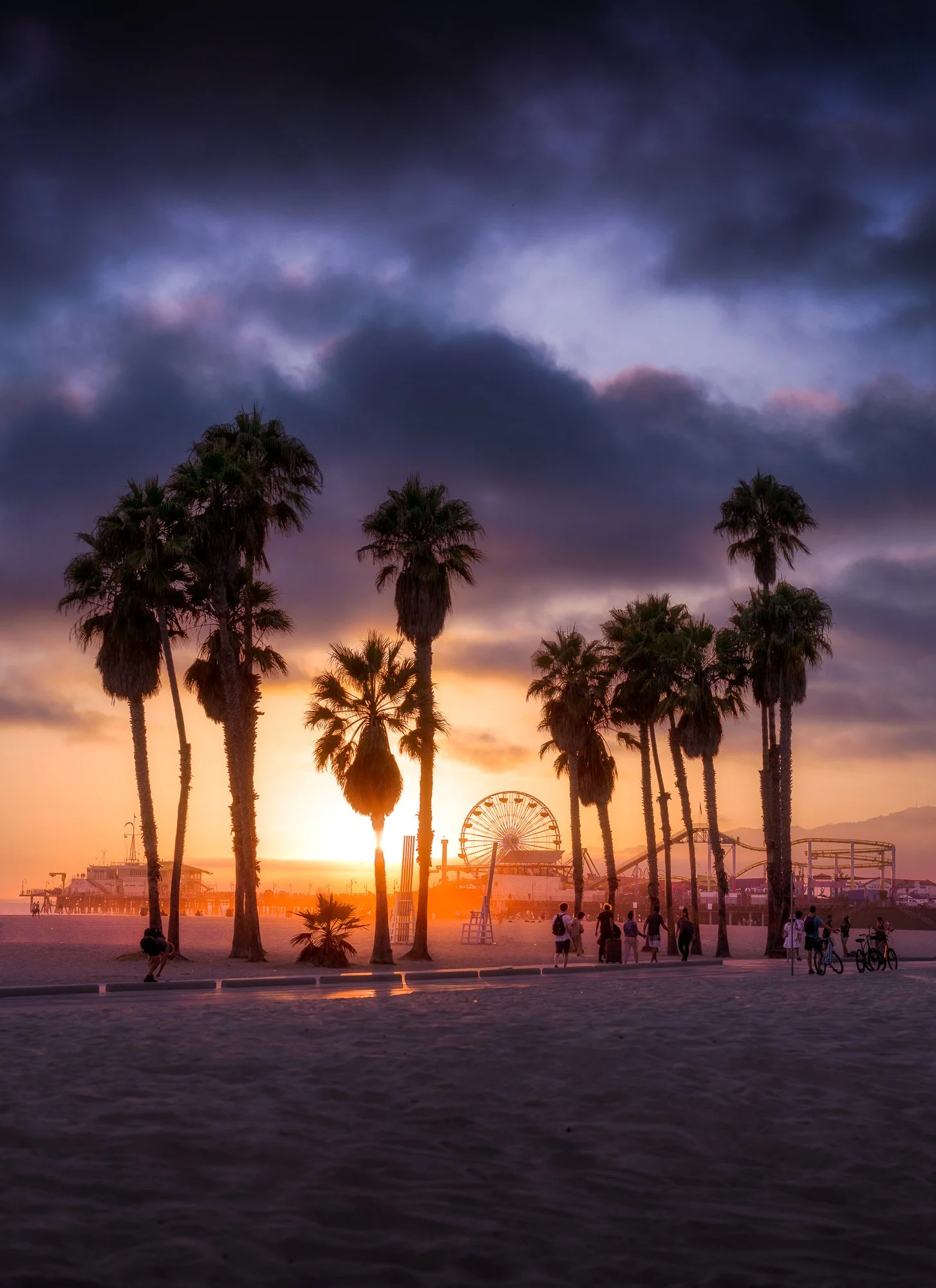 Sunset at a beach with palm trees, a ferris wheel, and people walking and biking.