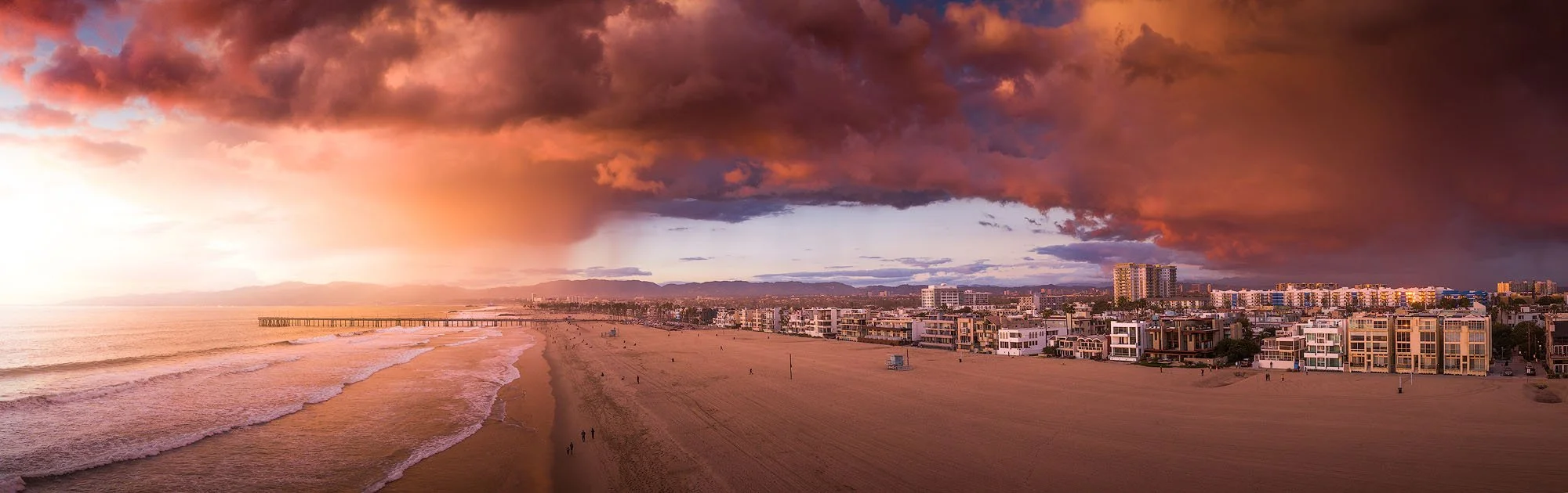 Beachside cityscape during a dramatic sunset with dark-colored clouds overhead and buildings along the shoreline.