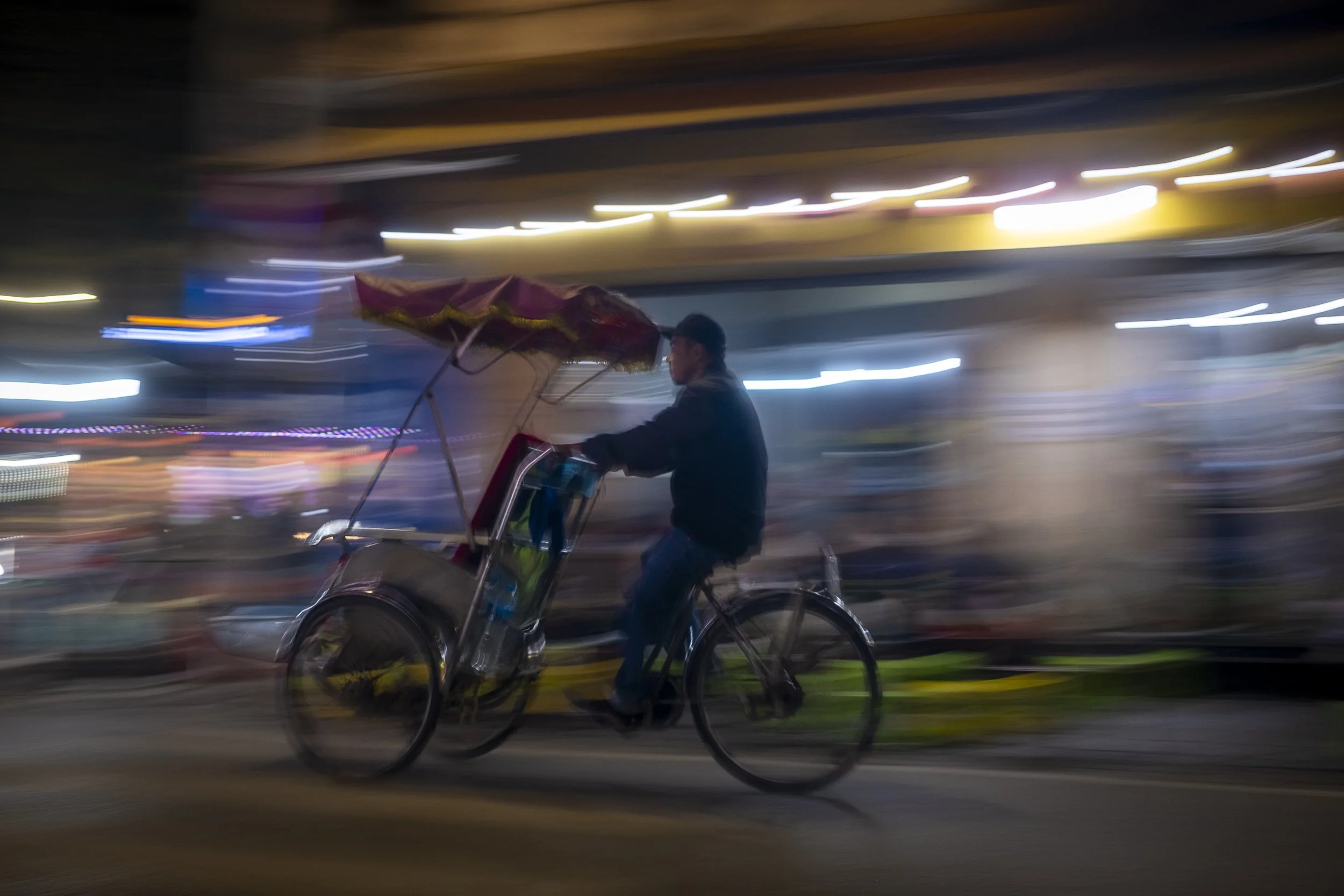A man riding a bicycle with a canopy attached, moving through a city street at night, with colorful lights creating a blurred background.