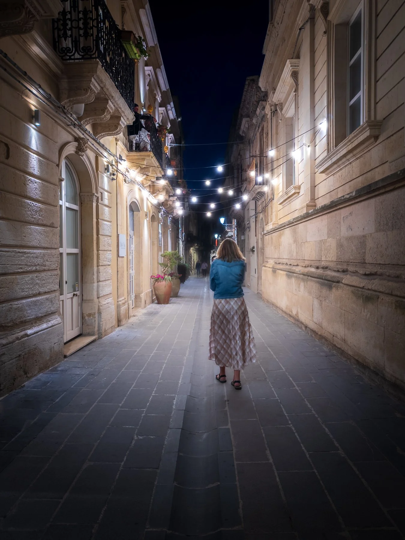 A woman walking on a lit cobblestone street at night, flanked by historic buildings with balconies and potted plants.