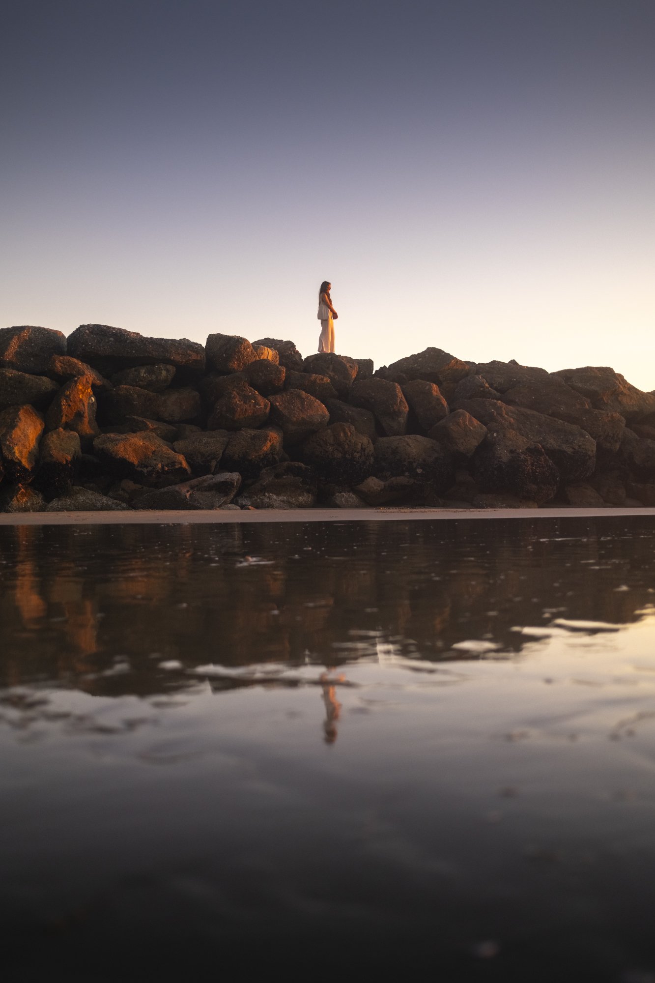 A woman standing on large rocks near the water during sunset, with her reflection visible in the calm water below.
