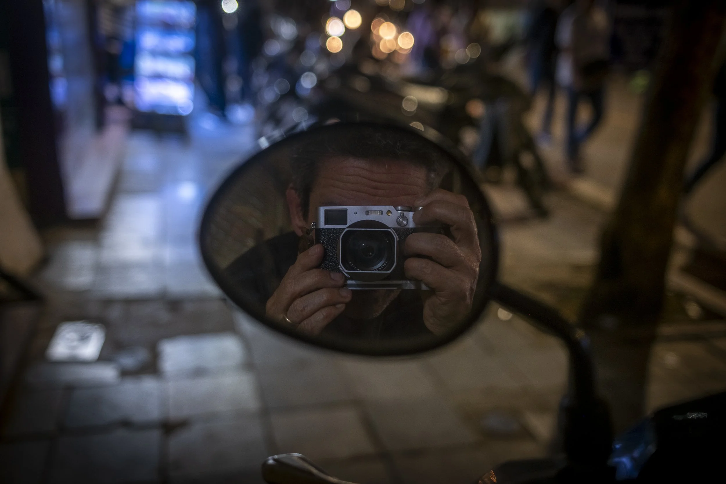 A person taking a photo of themselves with a camera in the motorcycle's rearview mirror inside a dimly lit room or garage.