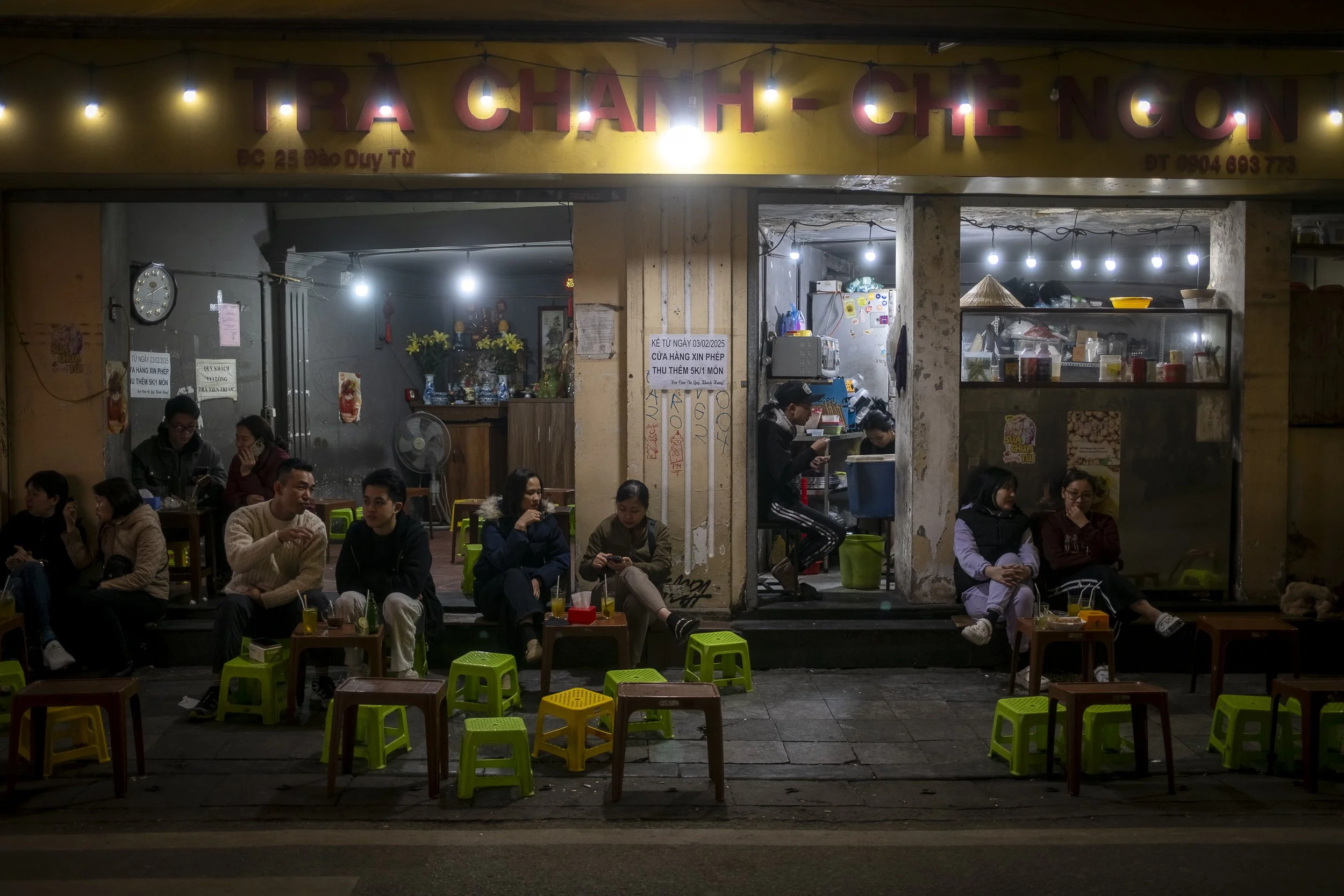 Night scene of a small Vietnamese restaurant with customers sitting outside on small plastic stools and tables, some eating and chatting, with the restaurant's bright yellow sign visible above and interior lighting illuminating the scene.
