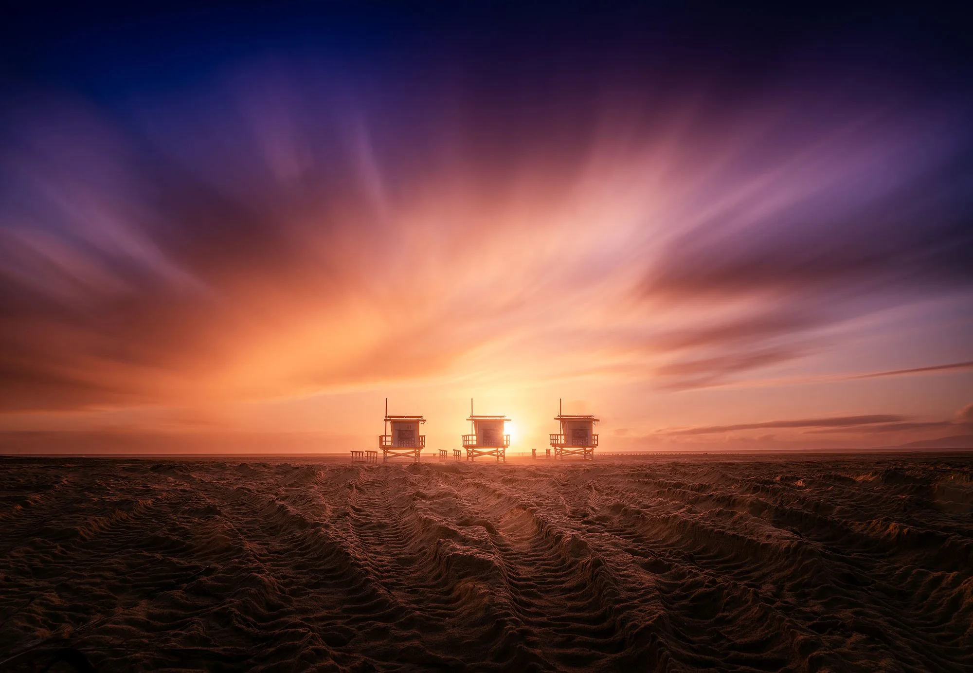 Three lifeguard towers on a sandy beach during a colorful sunset with purple, orange, and pink sky.