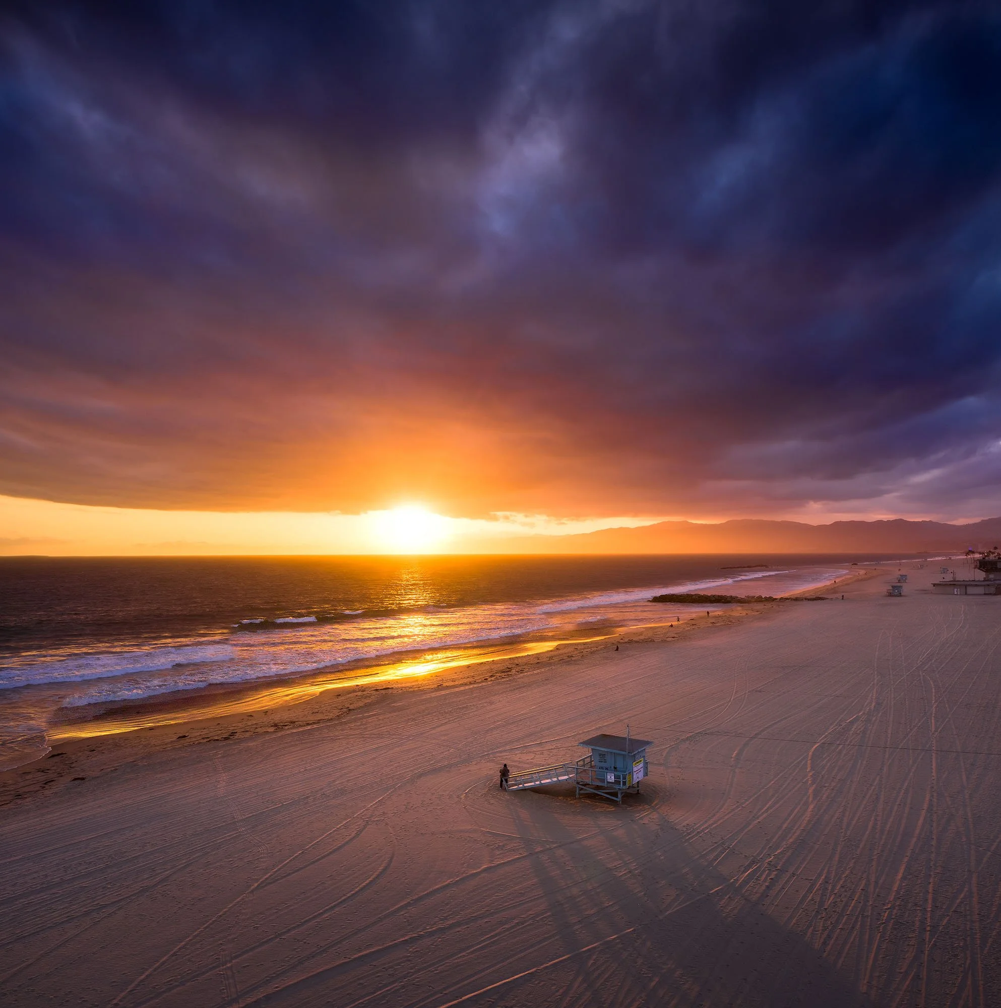 Sunset over a sandy beach with dark clouds overhead and a lifeguard tower in the foreground.