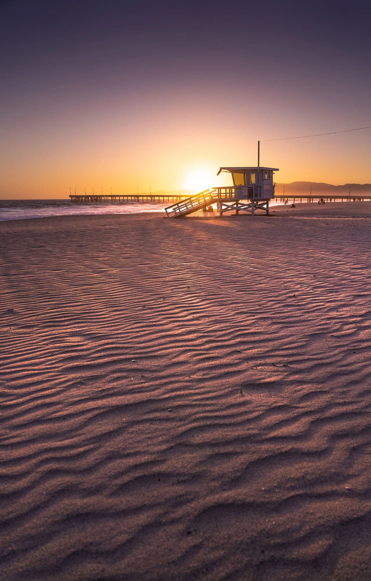 Sunset over a sandy beach with a lifeguard tower and a pier in the background.