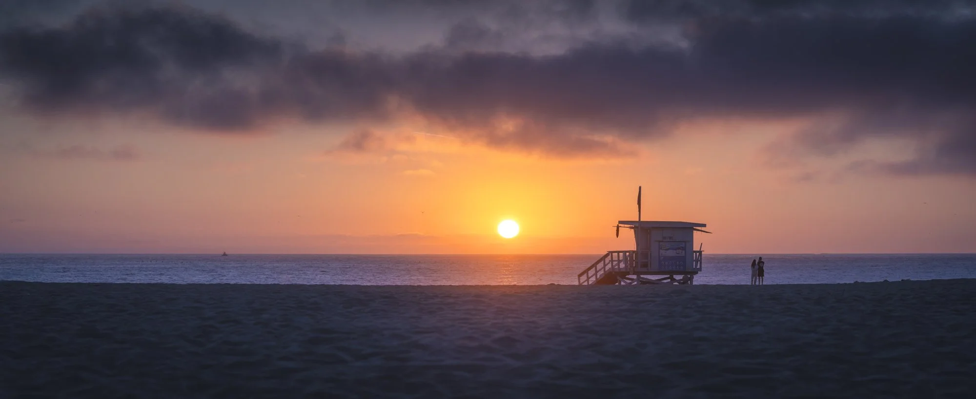 Sunset over the ocean with a lifeguard station and two people on the beach.