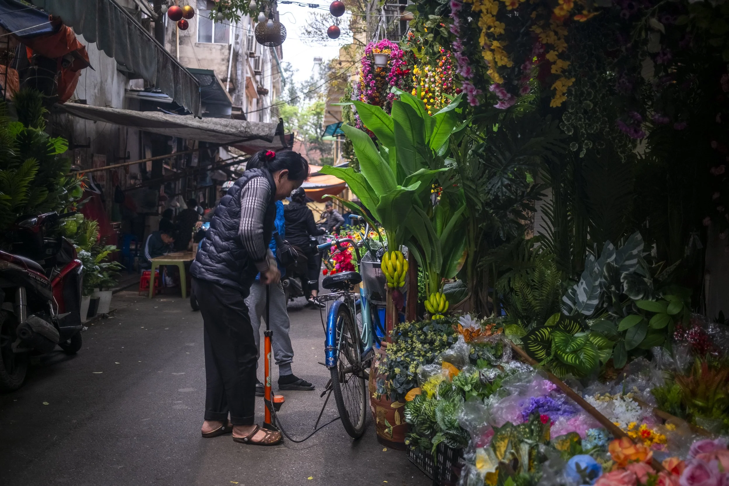 A woman checks her bicycle on a narrow street with flower stands on the side, featuring banana plants, colorful flowers, and greenery in an urban setting.