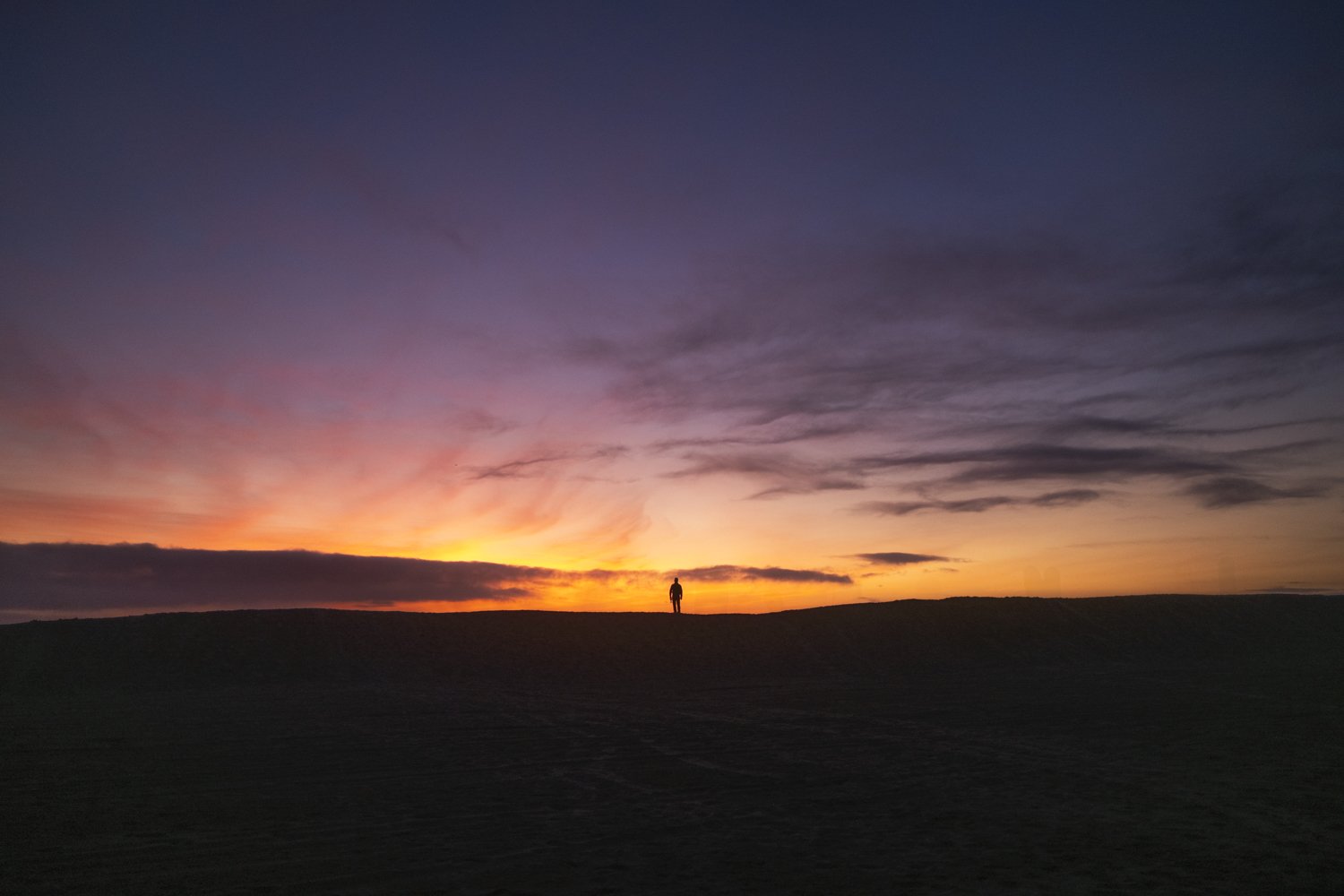 A person standing on a hill during sunset with a colorful sky featuring orange, pink, purple, and blue hues and scattered clouds.