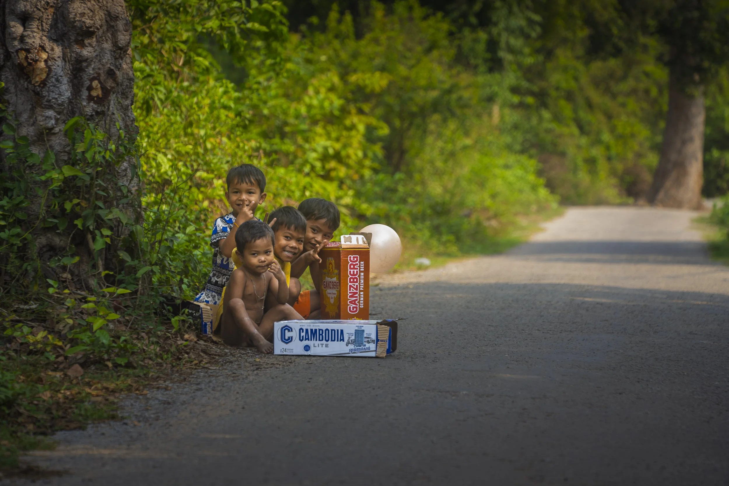 Group of five children sitting on the side of a rural road with trees in the background, surrounded by cardboard boxes and a plastic bottle, smiling and making gestures.