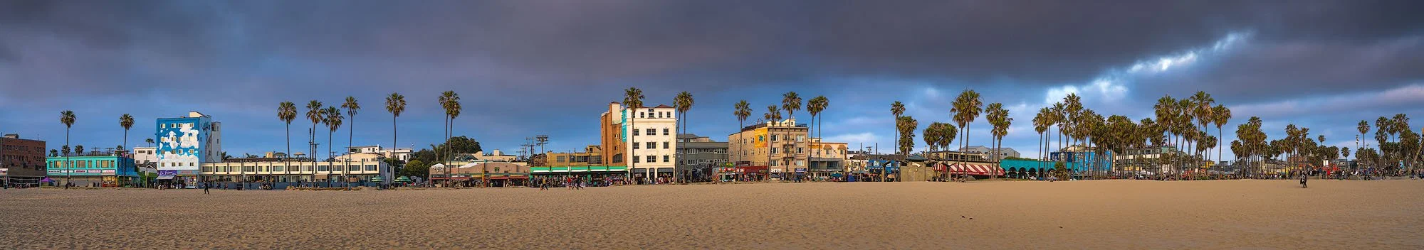 Panoramic view of Venice Beach boardwalk, with colorful buildings, palm trees, and shops, under a stormy sky.