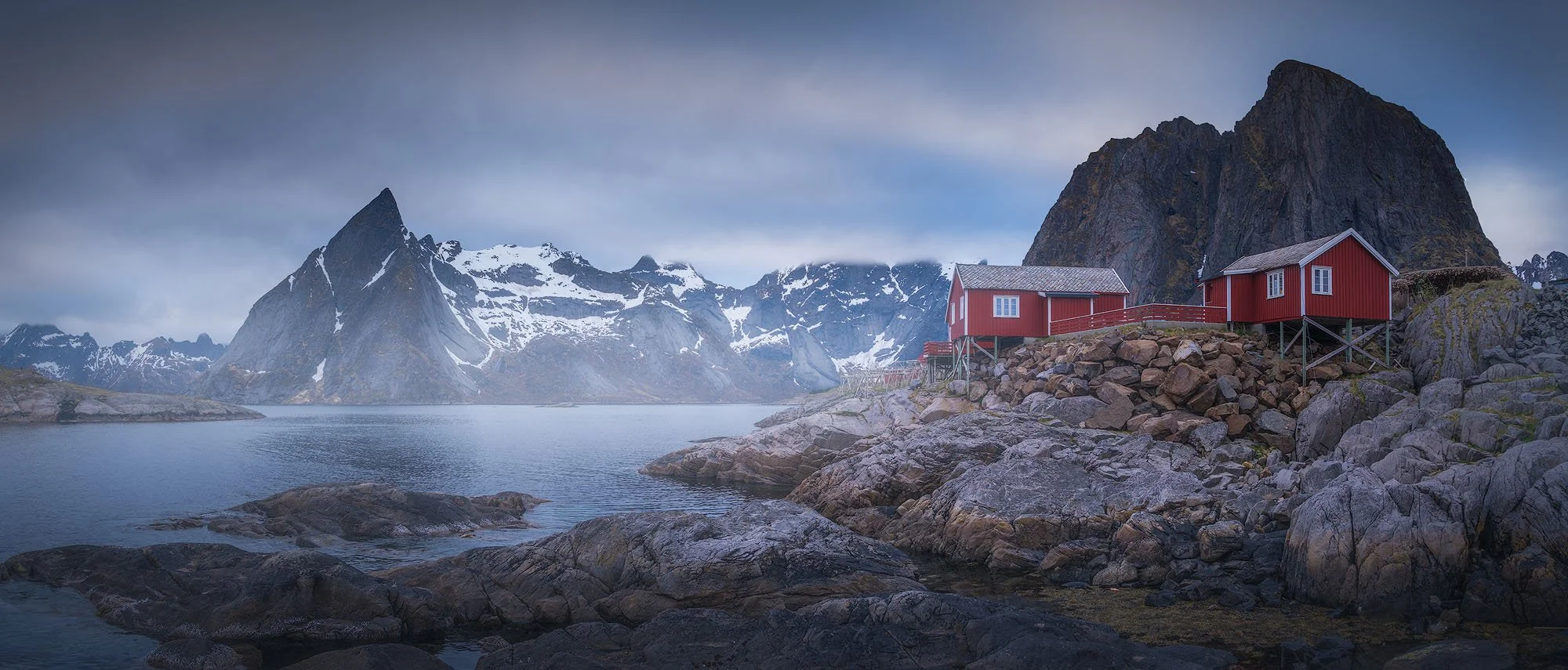 Red cabins on rocky shore with mountains and snow in the background.
